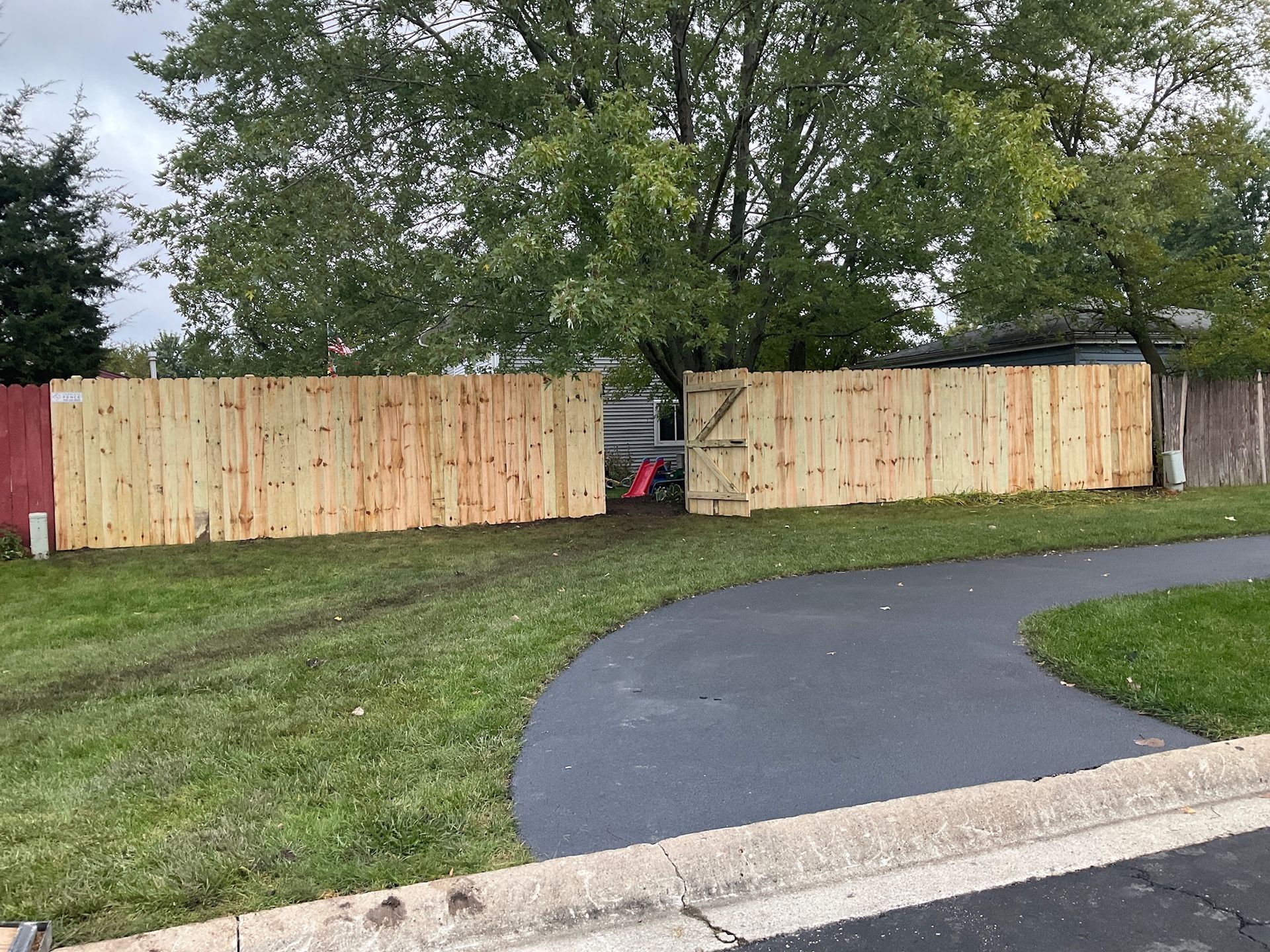 Wooden fence with gate opening onto a driveway, green grass, and trees in the background.