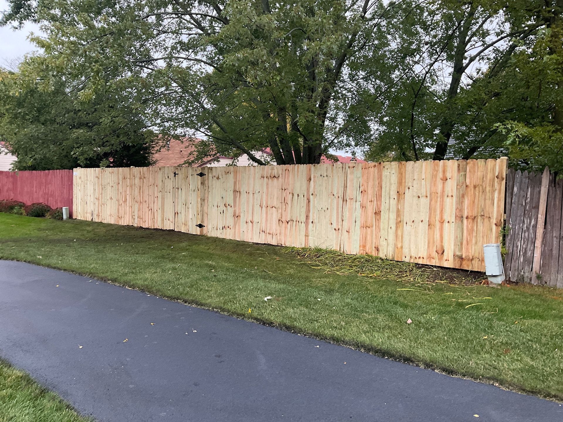 Wooden fence along a paved path, transitioning from red to natural wood, with grass and trees in the background.
