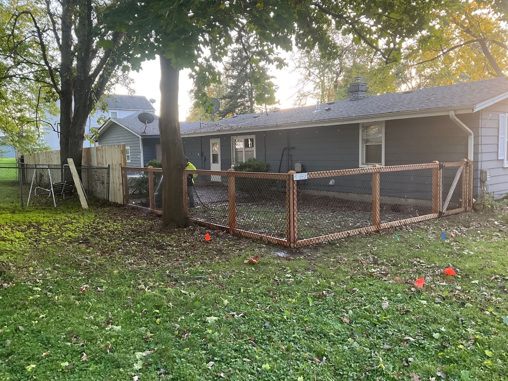 A backyard with a wooden fence enclosing an area near a gray house; autumn leaves on the grass.