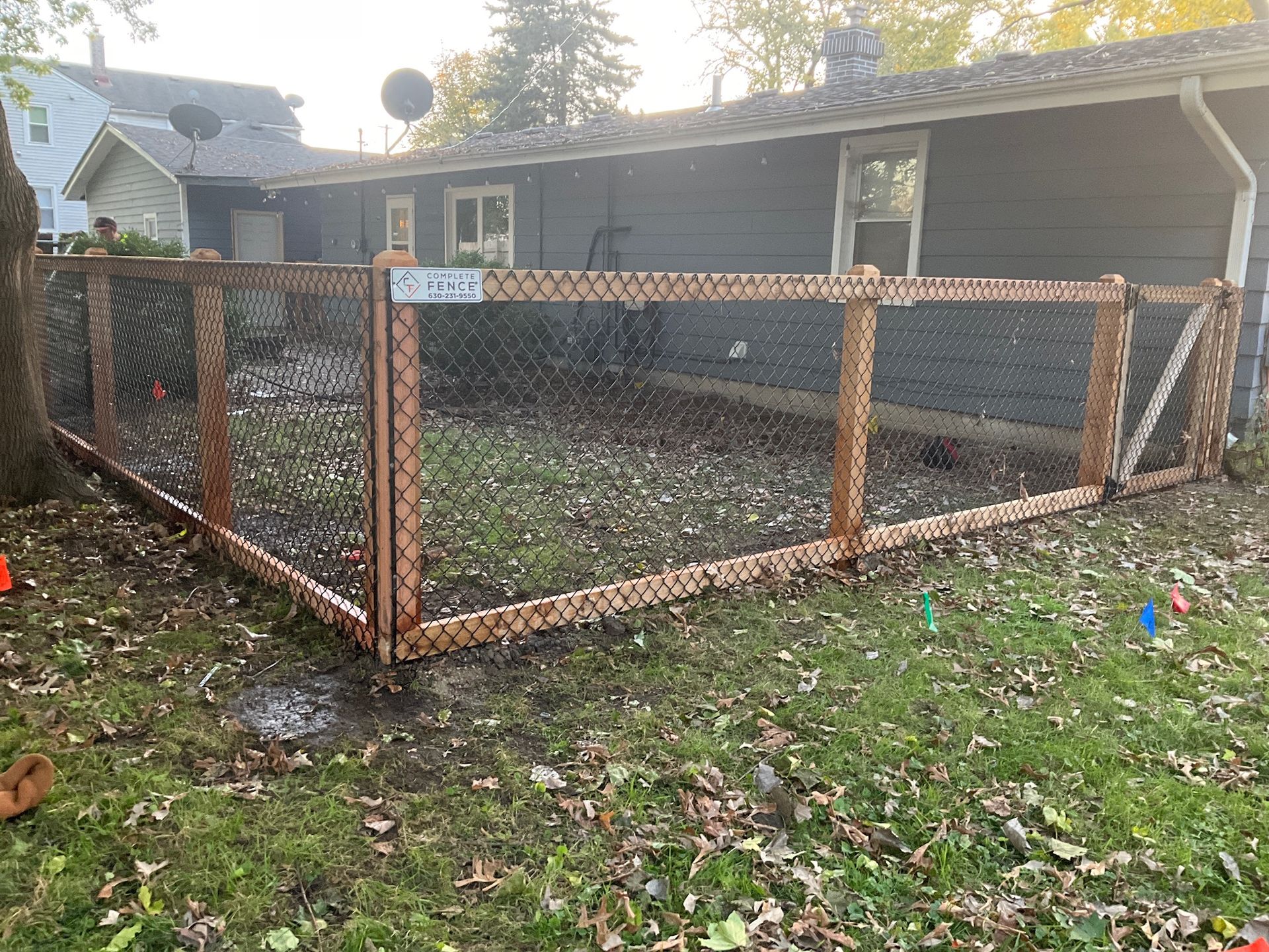A wooden and wire fence surrounds a grassy backyard, adjacent to a gray house.