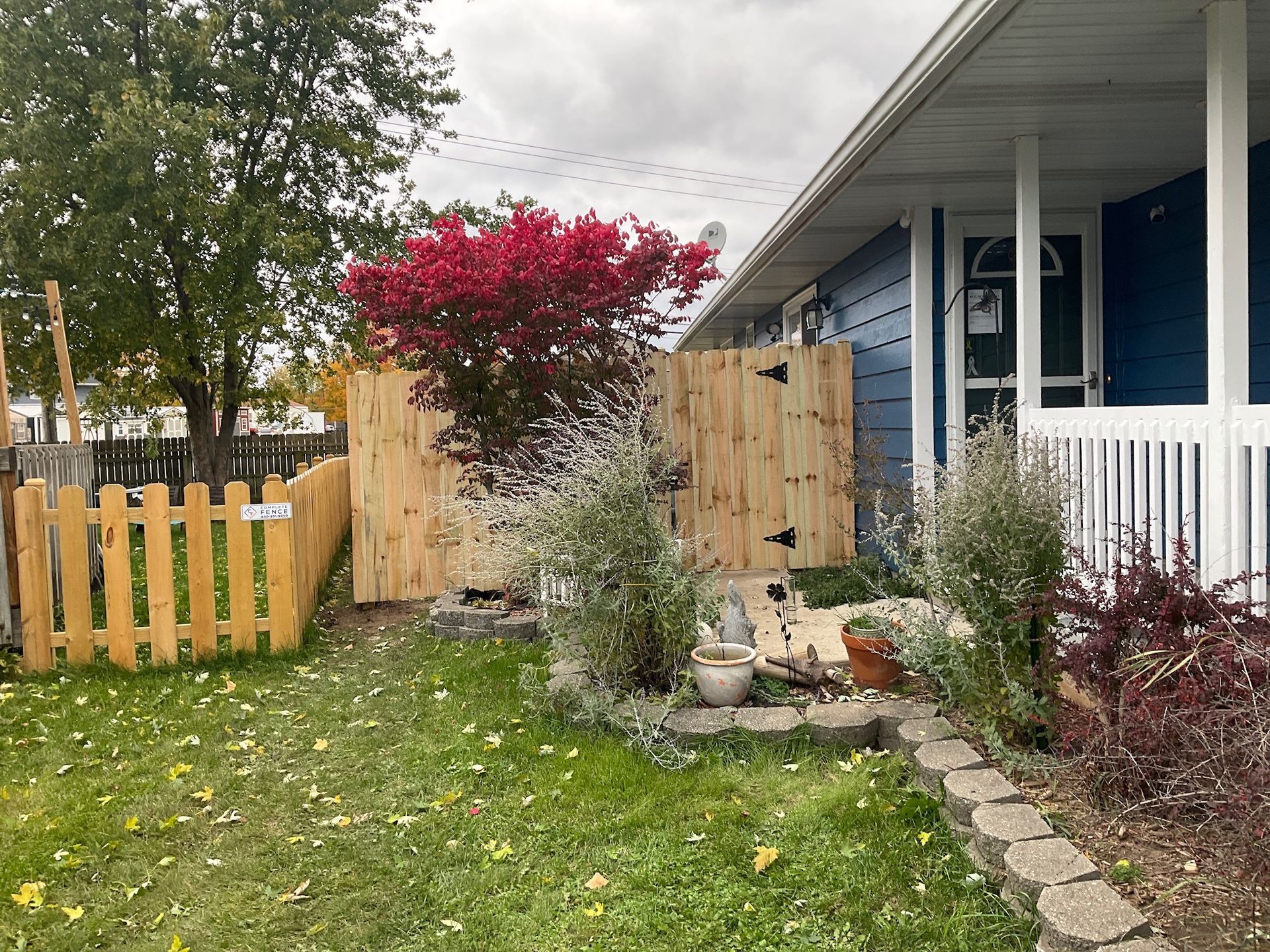 Wooden fences surround a front yard with a blue house. Green grass, a red tree, and bushes are visible.