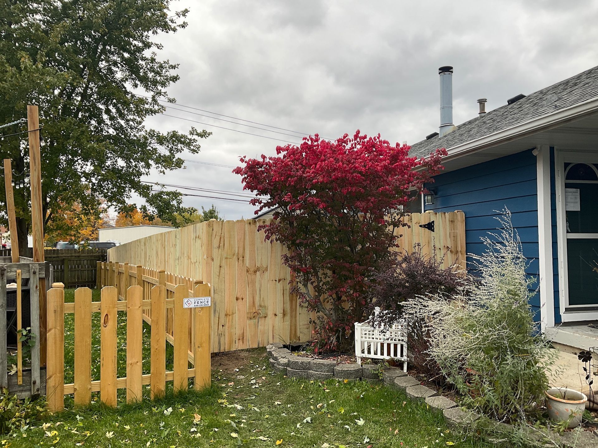 Wooden fence in front of a blue house with a red-leaved bush and a small wooden gate. Cloudy sky.
