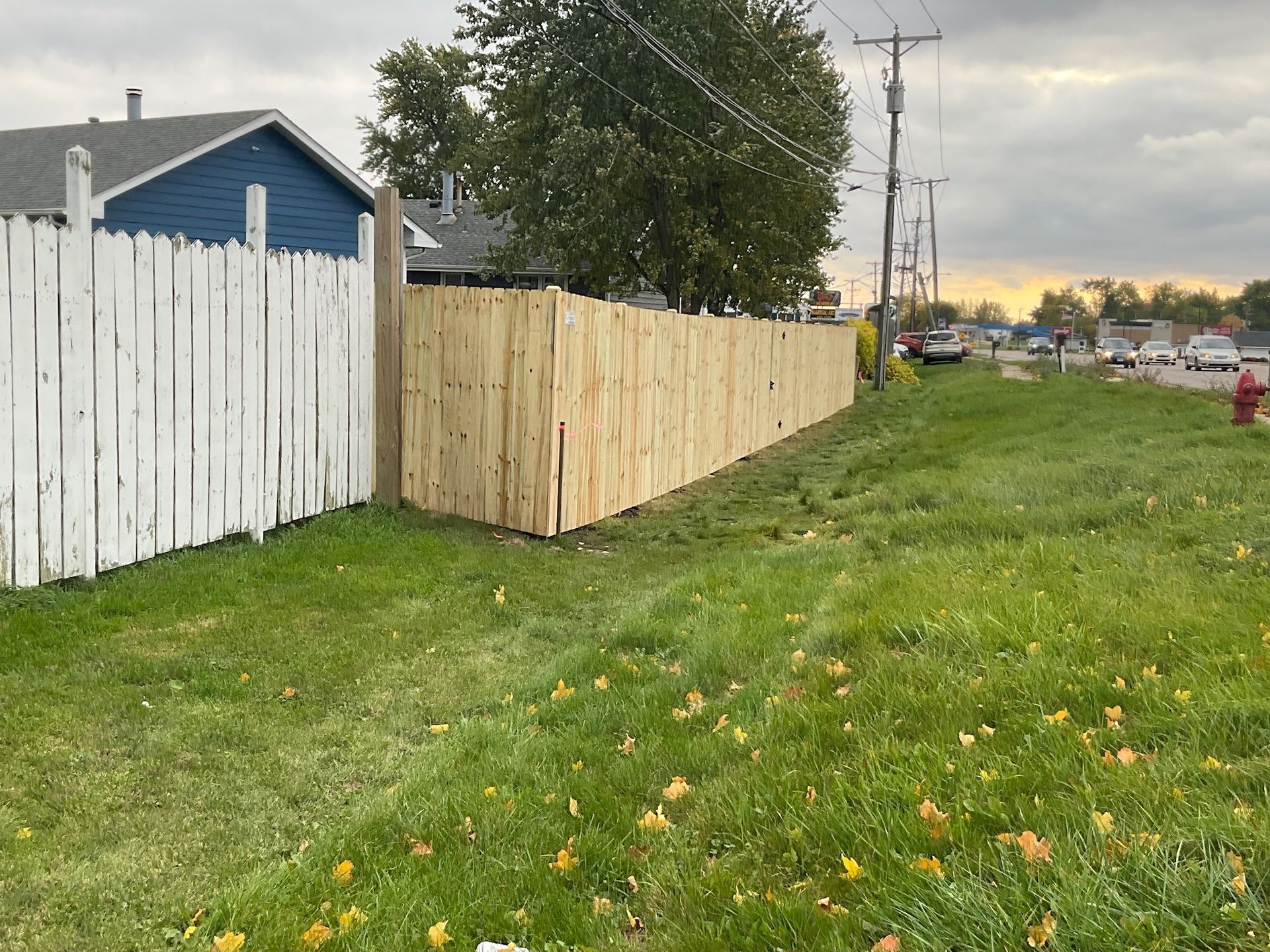 A wooden fence runs along a grassy lawn next to a road, with a blue house visible.