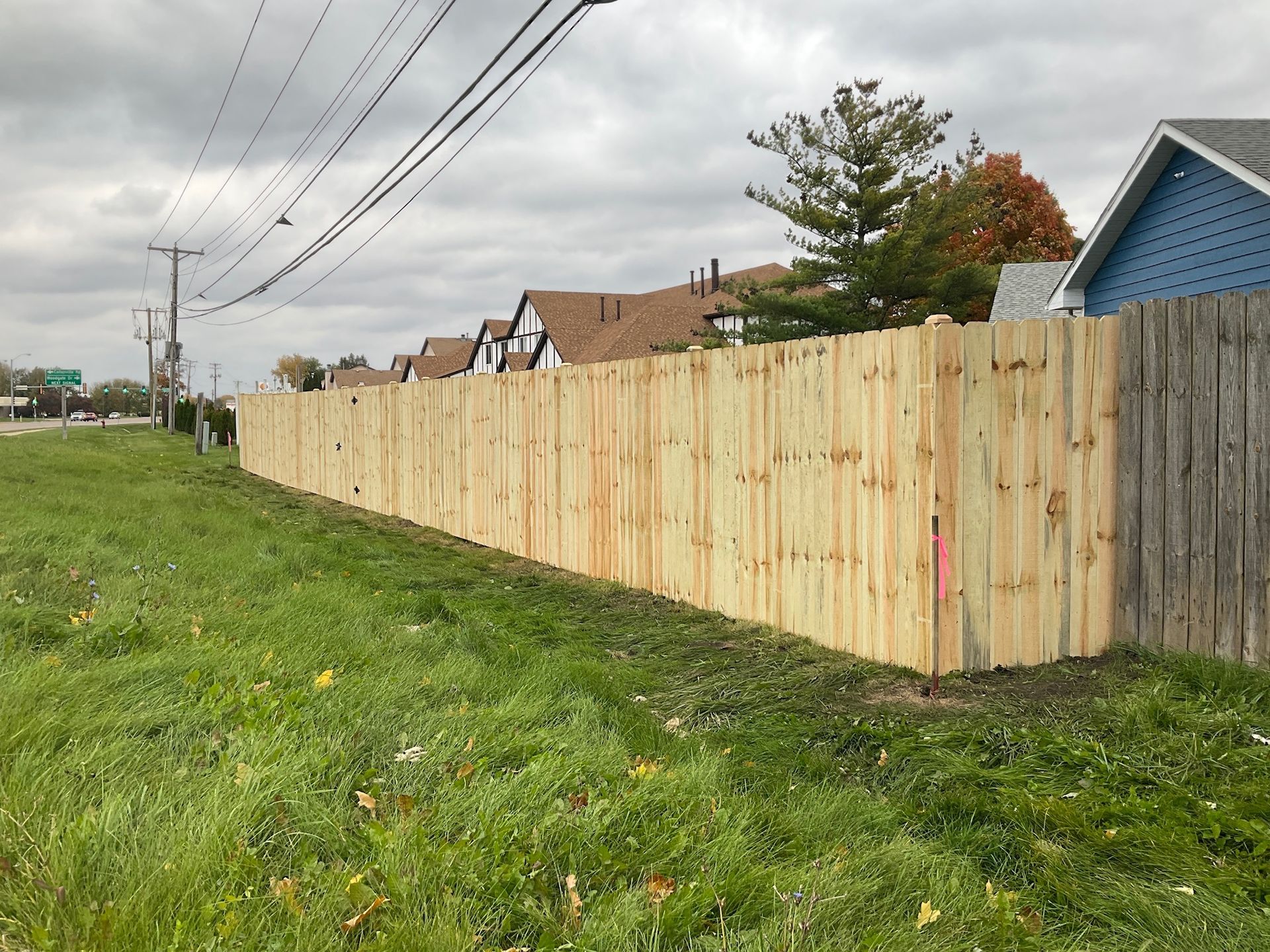 Wooden fence bordering a grassy area next to a road, houses in the background under a cloudy sky.
