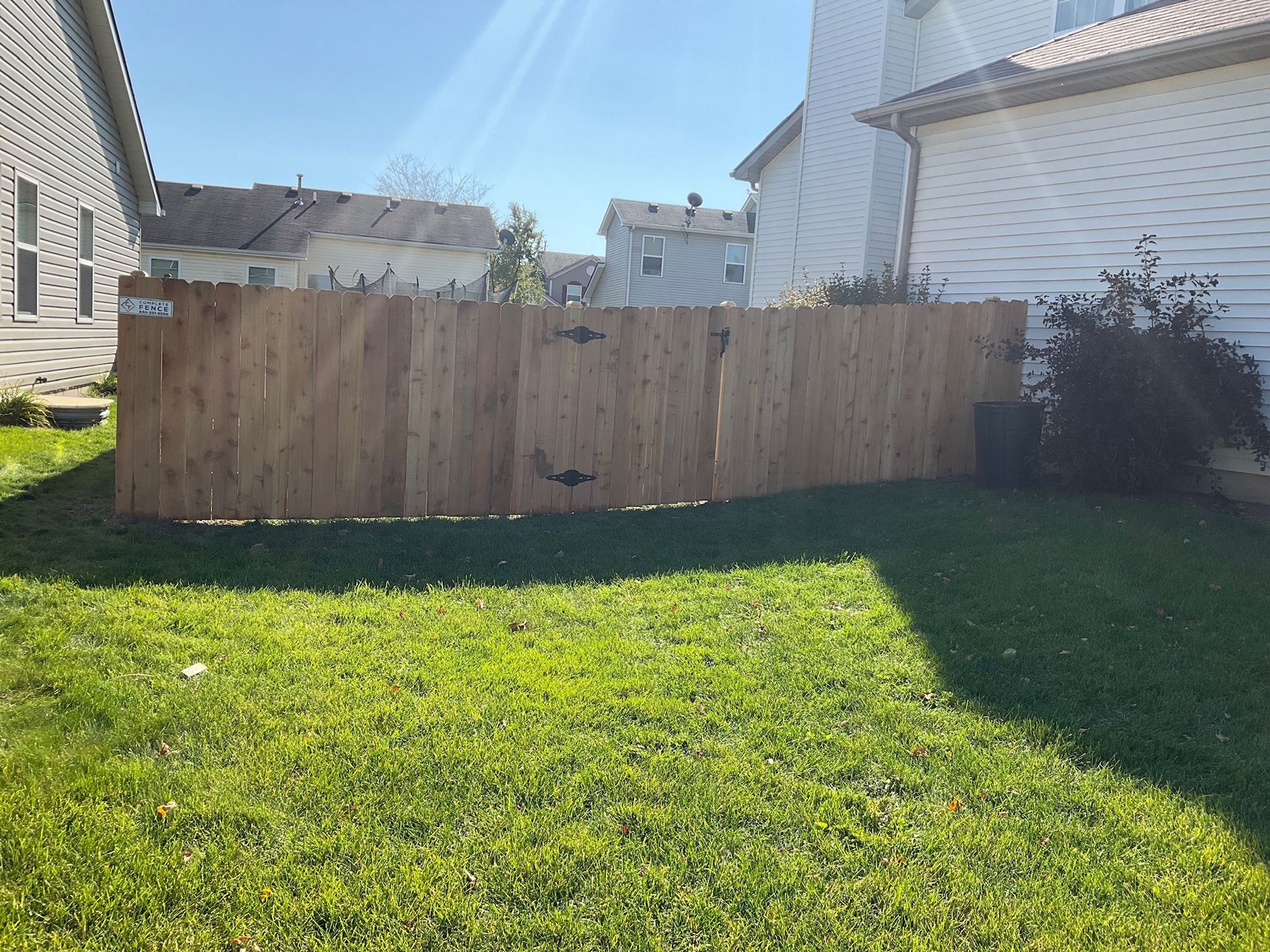 Wooden fence in a backyard with green grass and sunlight. Houses visible in the background.