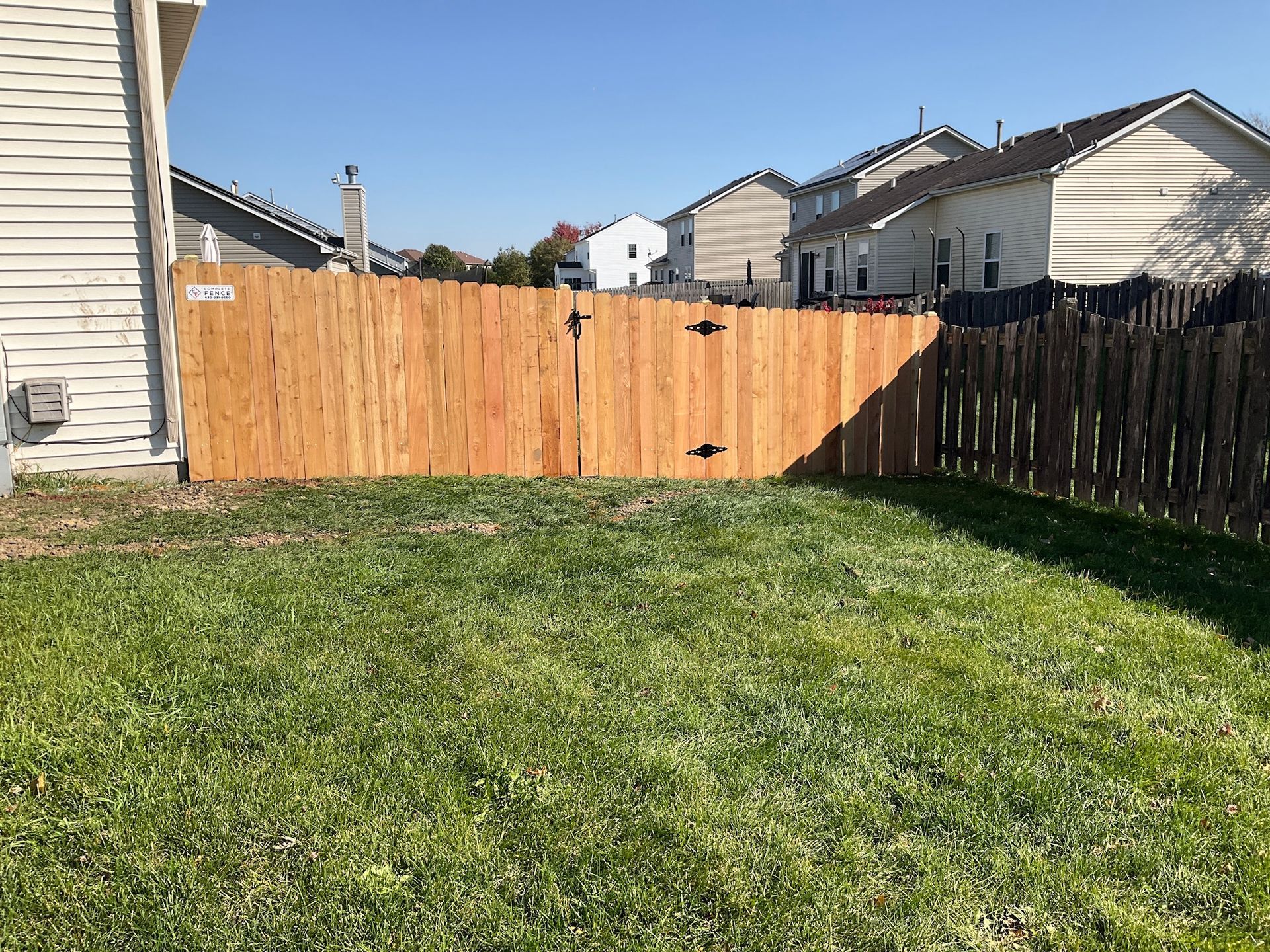 Wooden fence in a grassy backyard with houses in the background on a sunny day.