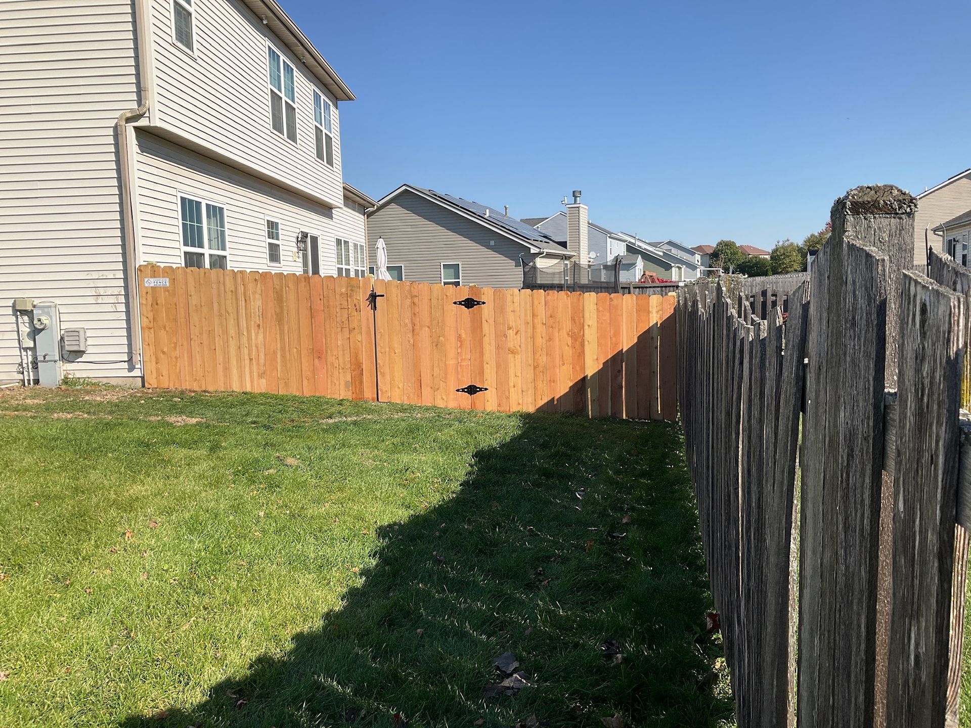 Backyard scene: new stained wood fence beside weathered fence. Sunny day, green grass, two-story houses.