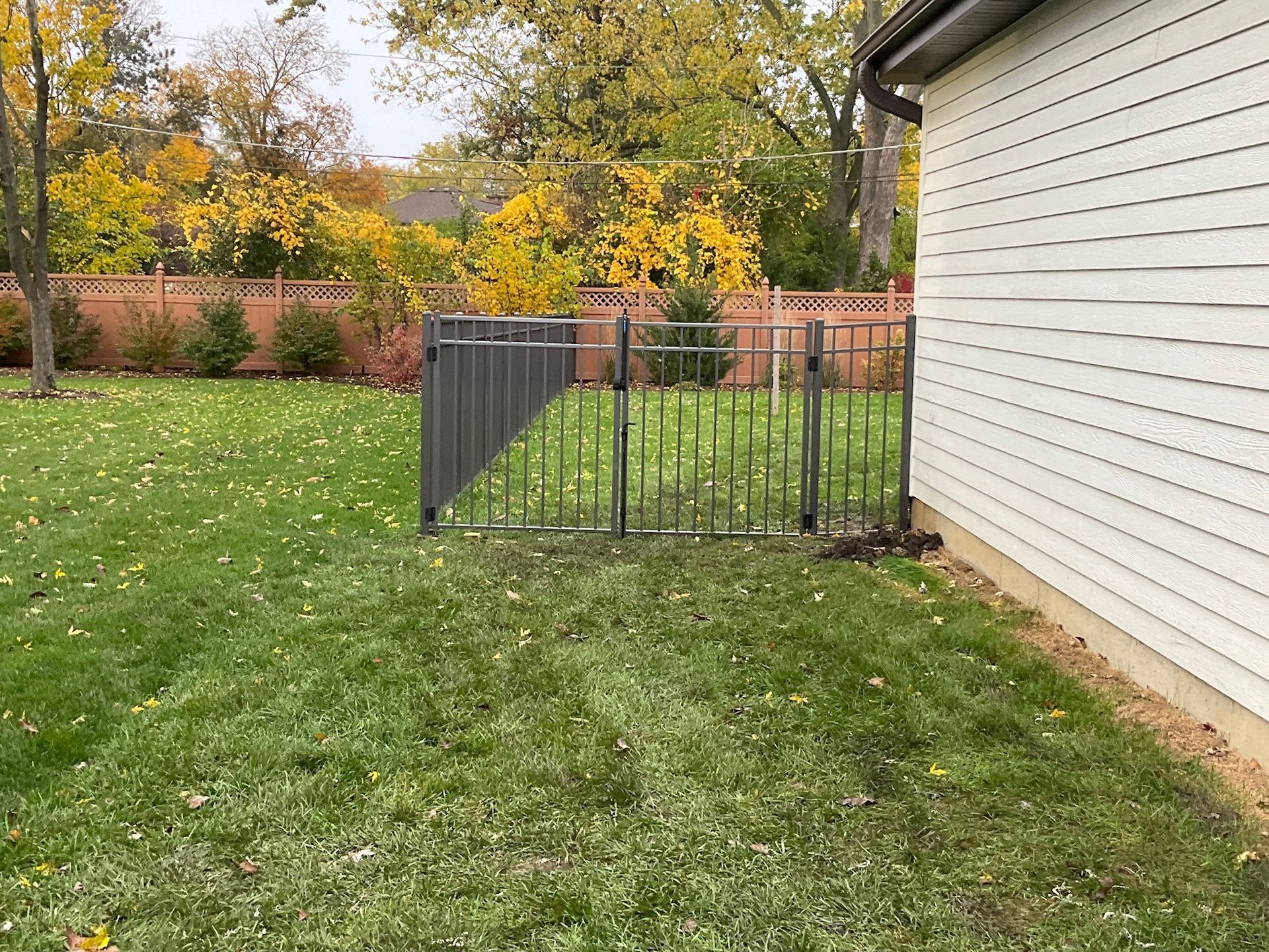 A gray metal gate in a green grassy yard next to a white house with an autumn backdrop.