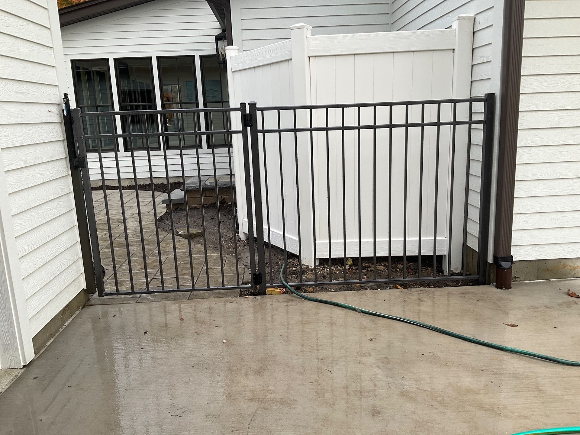 Black metal gate in front of white fence, on a concrete patio, with siding and a green hose.