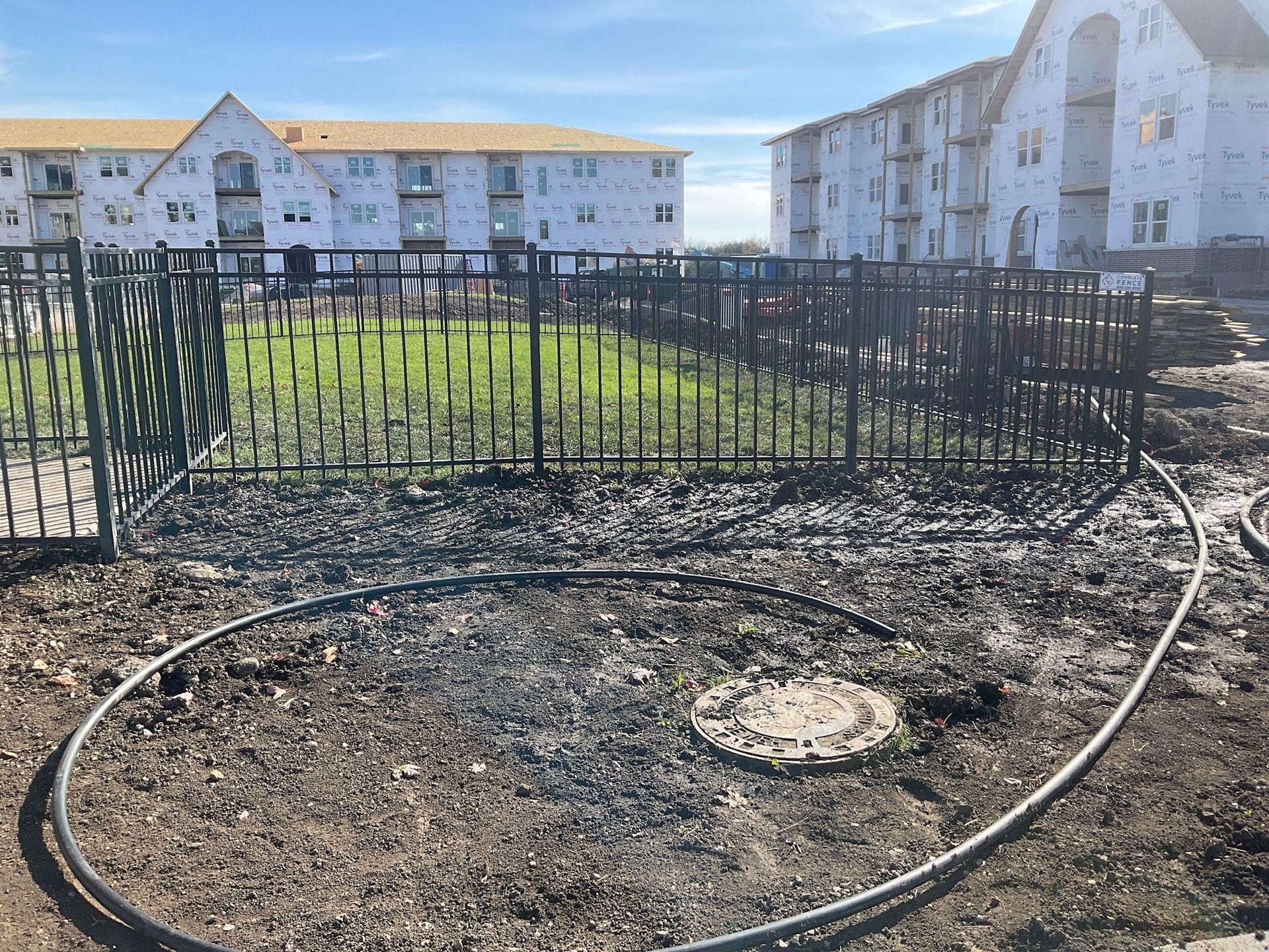 Fenced-in construction site with muddy ground, hose, and unfinished apartment buildings in the background.