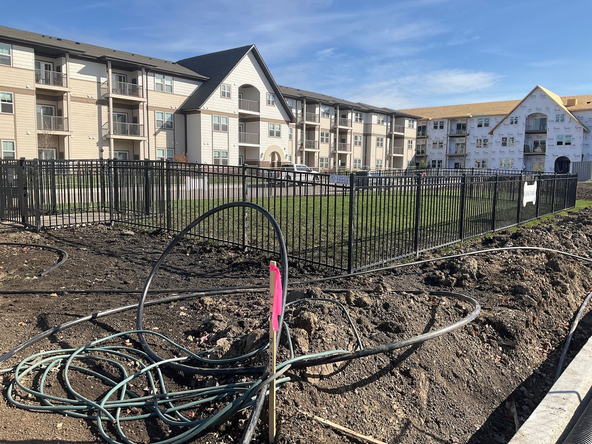 Construction site in front of apartment building; black fence encloses a green lawn. Wires and dirt in foreground.