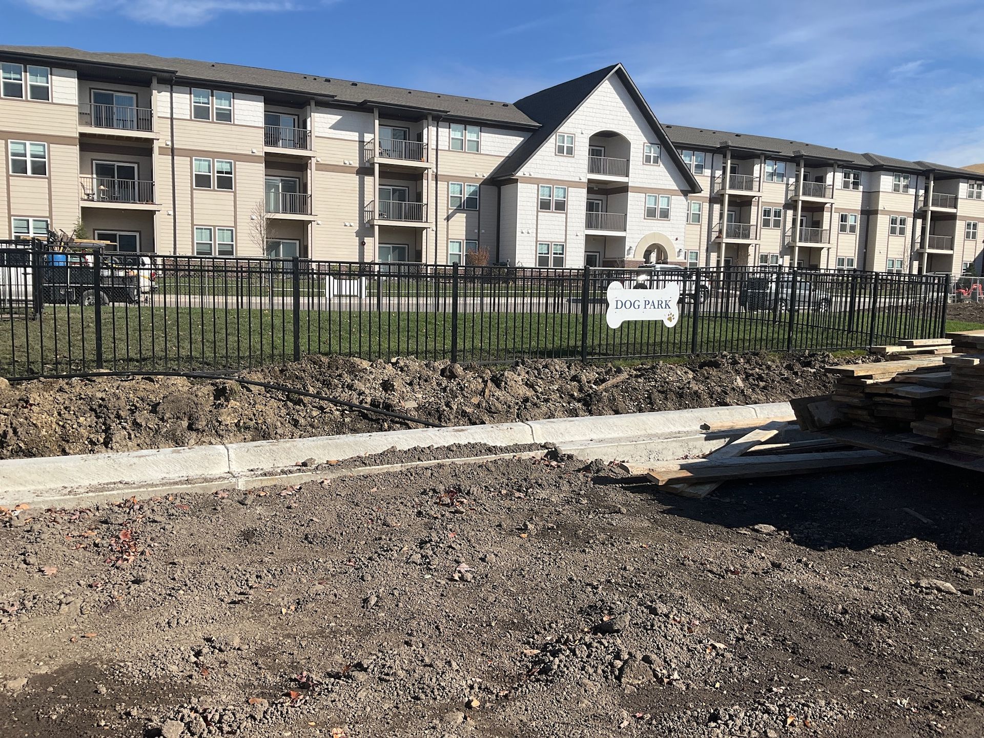 Construction site: a building with balconies behind a black fence. Dirt and gravel in the foreground.