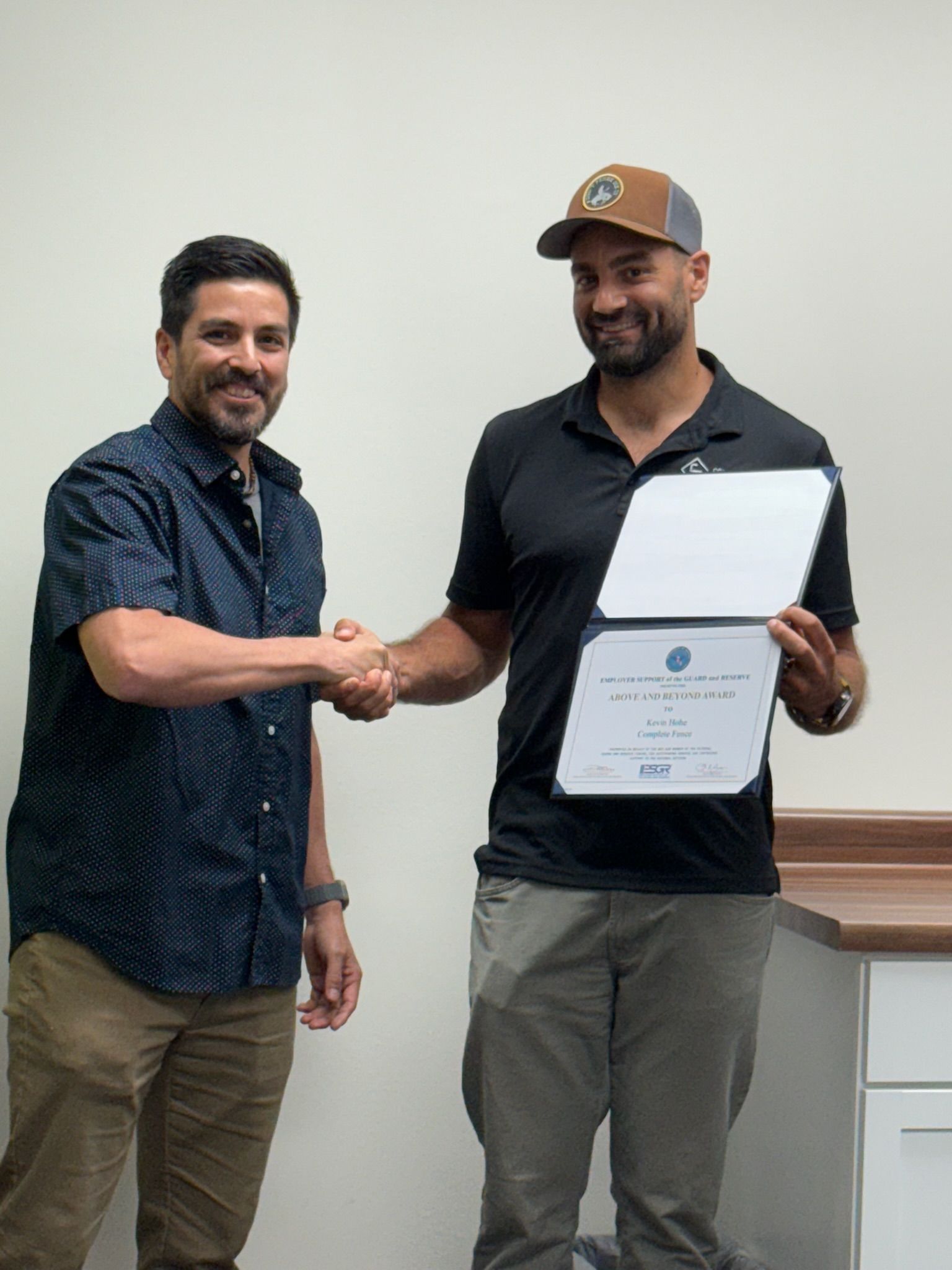Two men shake hands; one holds a certificate. Light-skinned room, tan pants, black shirt, and a brown cap.