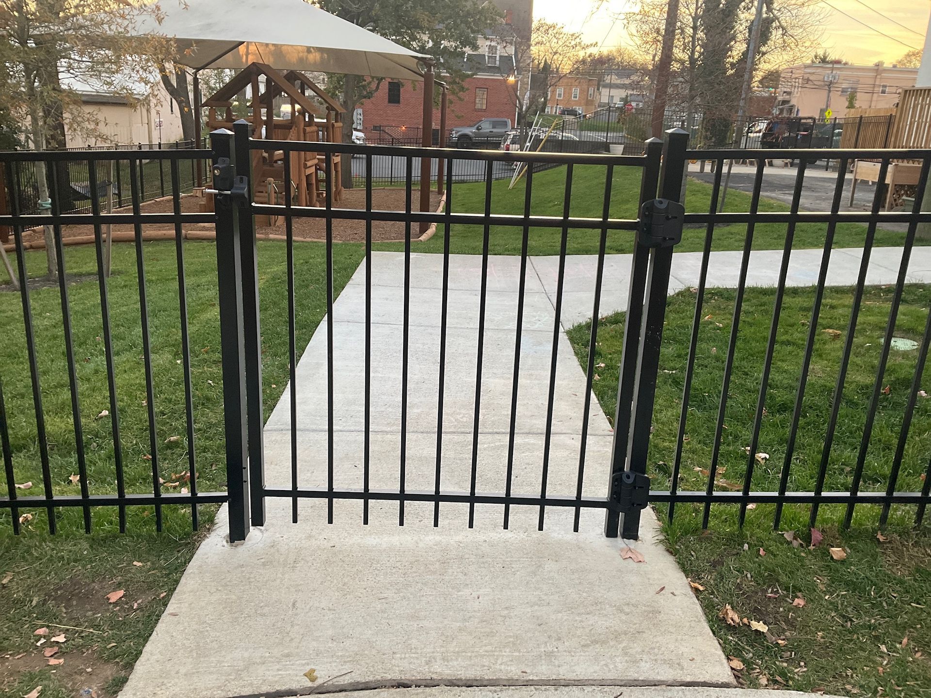 Black metal gate in a fenced yard, with a concrete pathway leading through it. Green grass surrounds.