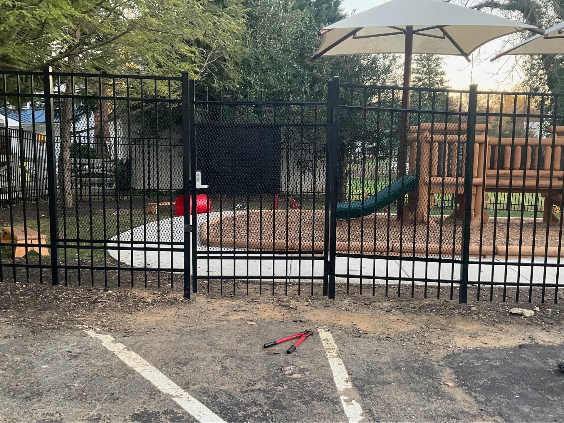 Black metal fence with gate, enclosing a playground with wood structure and umbrella.