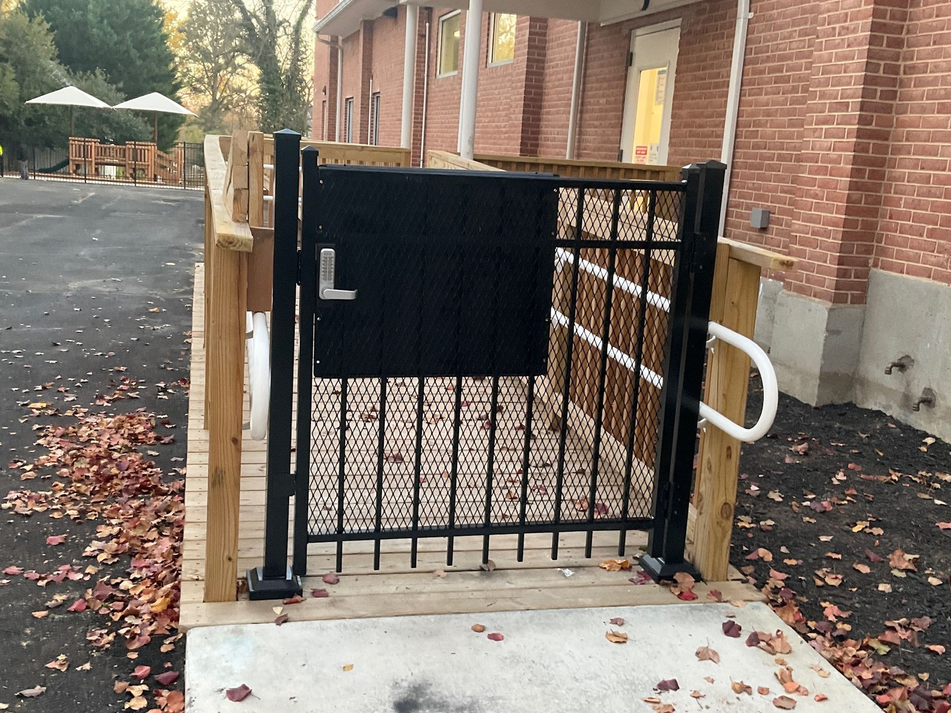 Black metal gate on wooden ramp with handrail; brick building in the background, leaves on ground.