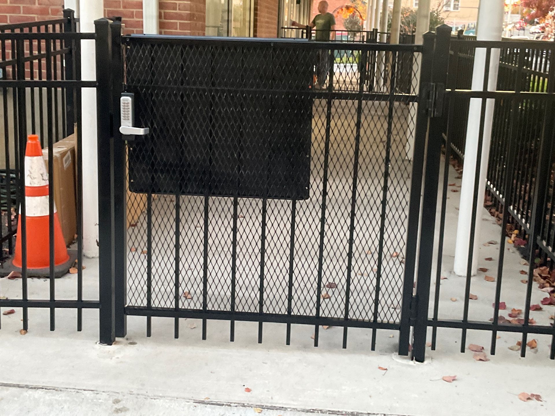 Black metal gate with a lock. The gate has a perforated metal panel. A traffic cone is to the left.