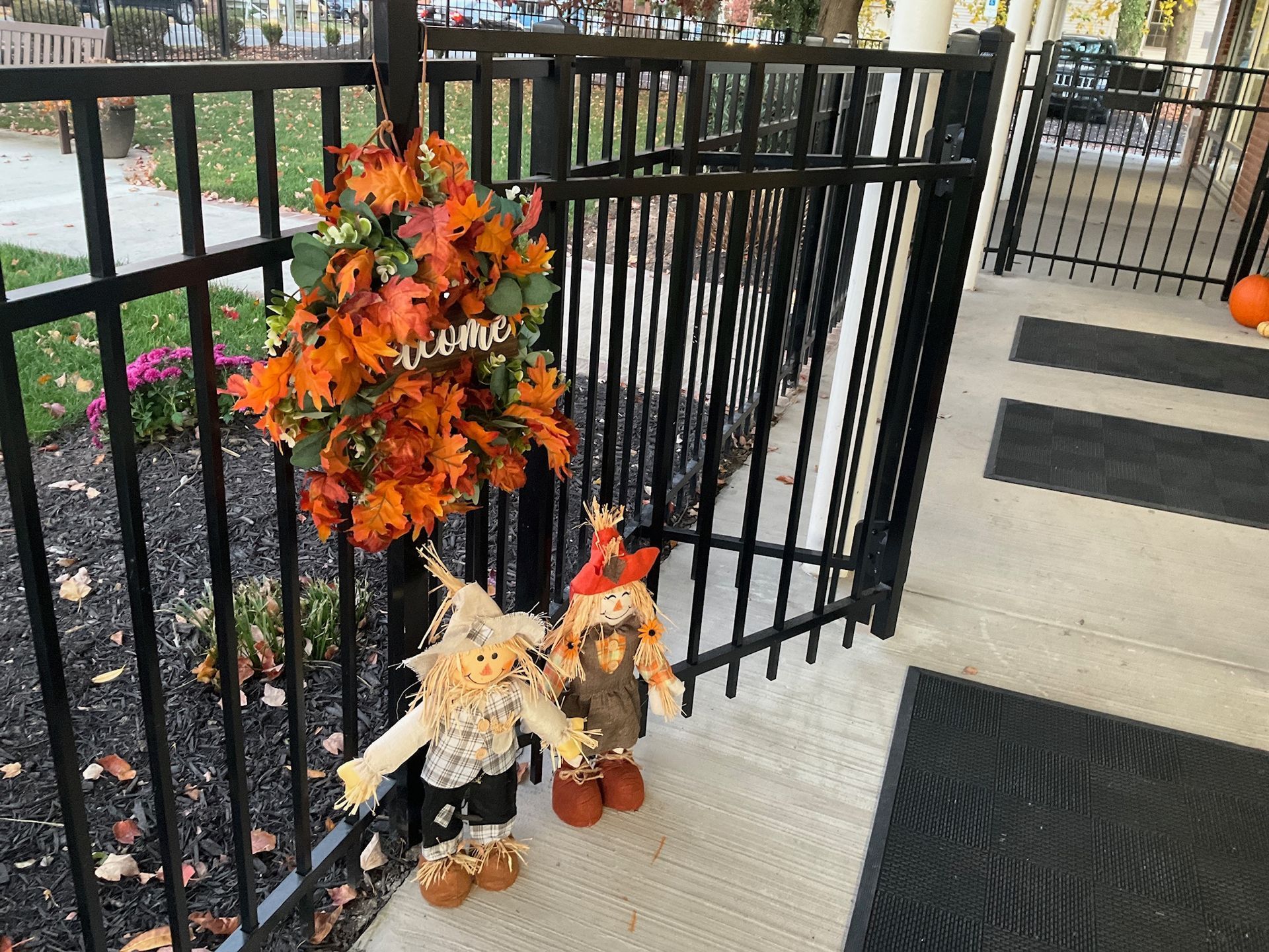 Fall decorations: wreath, scarecrows, on a porch with black railing.