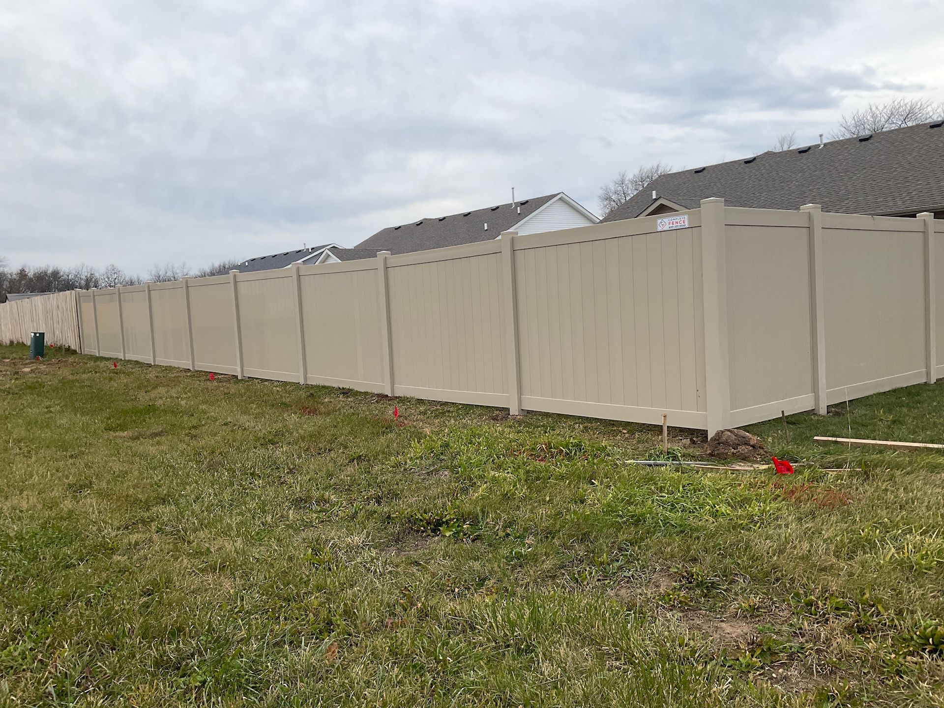 Tan vinyl fence in a grassy yard, houses in the background under a cloudy sky.
