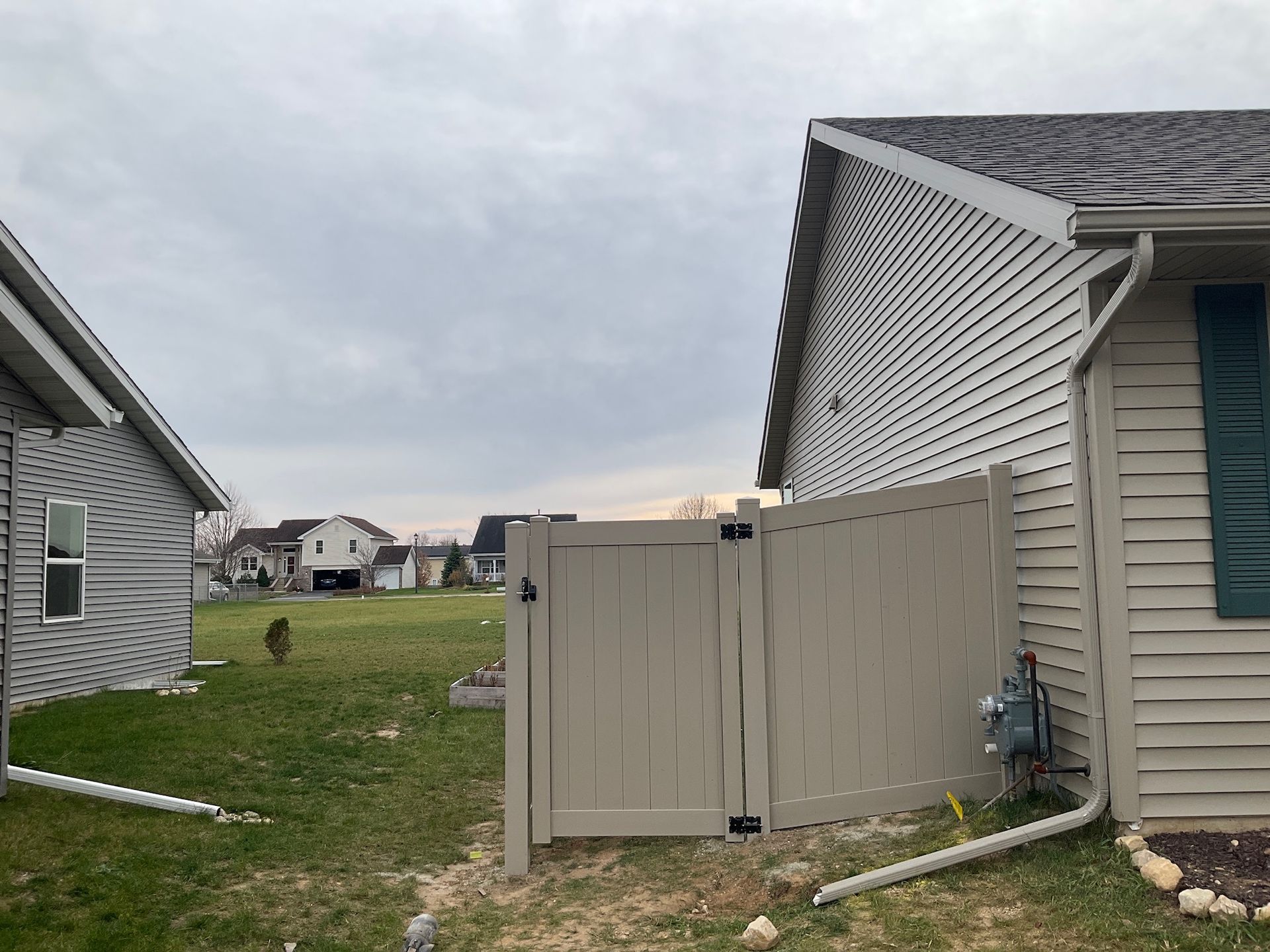 Tan vinyl gate between two houses on a grassy yard under a cloudy sky.