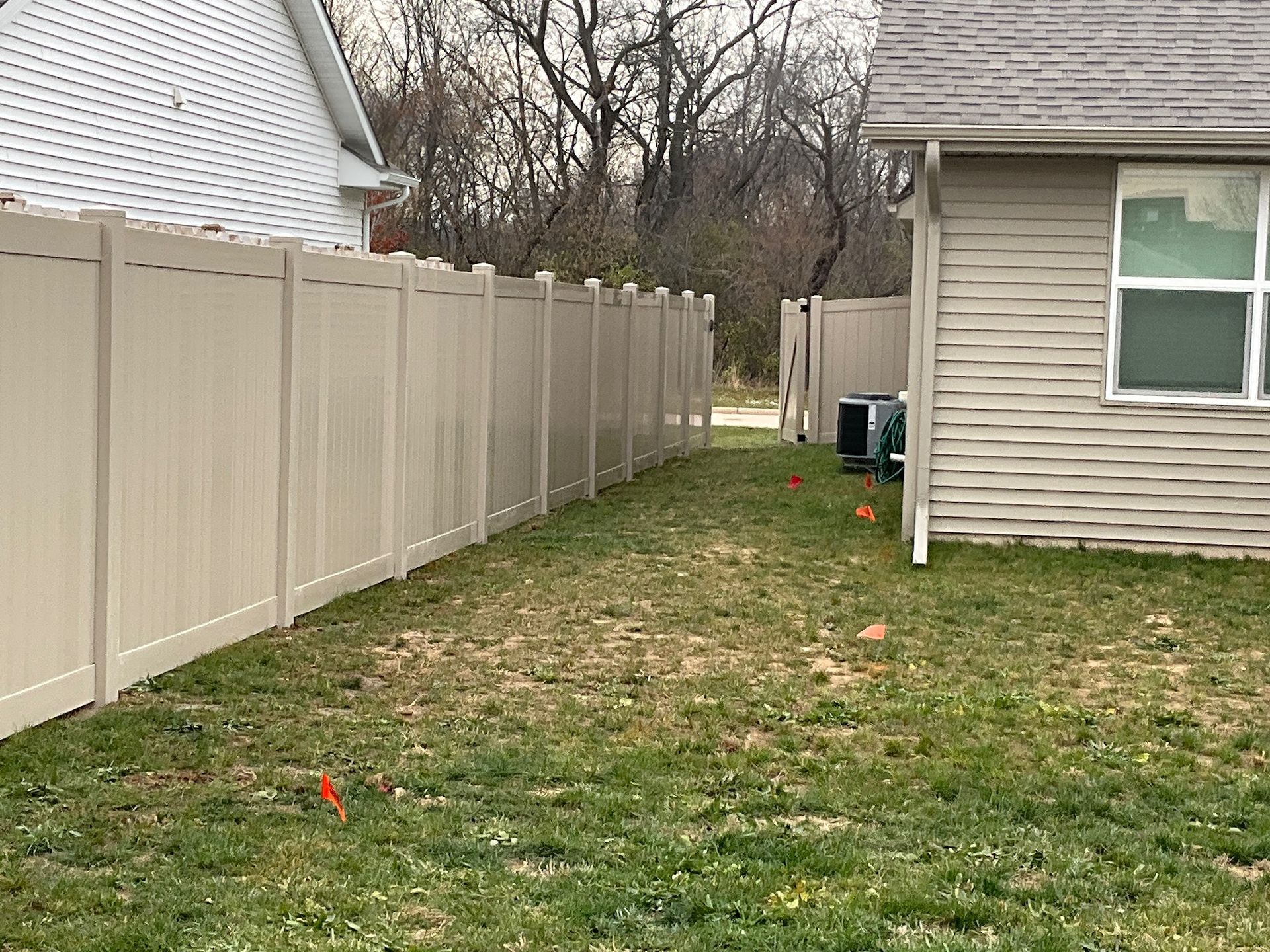 Tan vinyl fence surrounds a yard with patchy grass next to a house with beige siding.