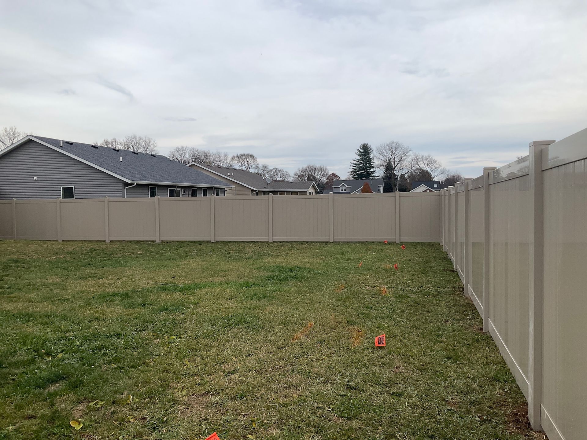 A tan vinyl privacy fence encloses a backyard with brown and green grass, on an overcast day.