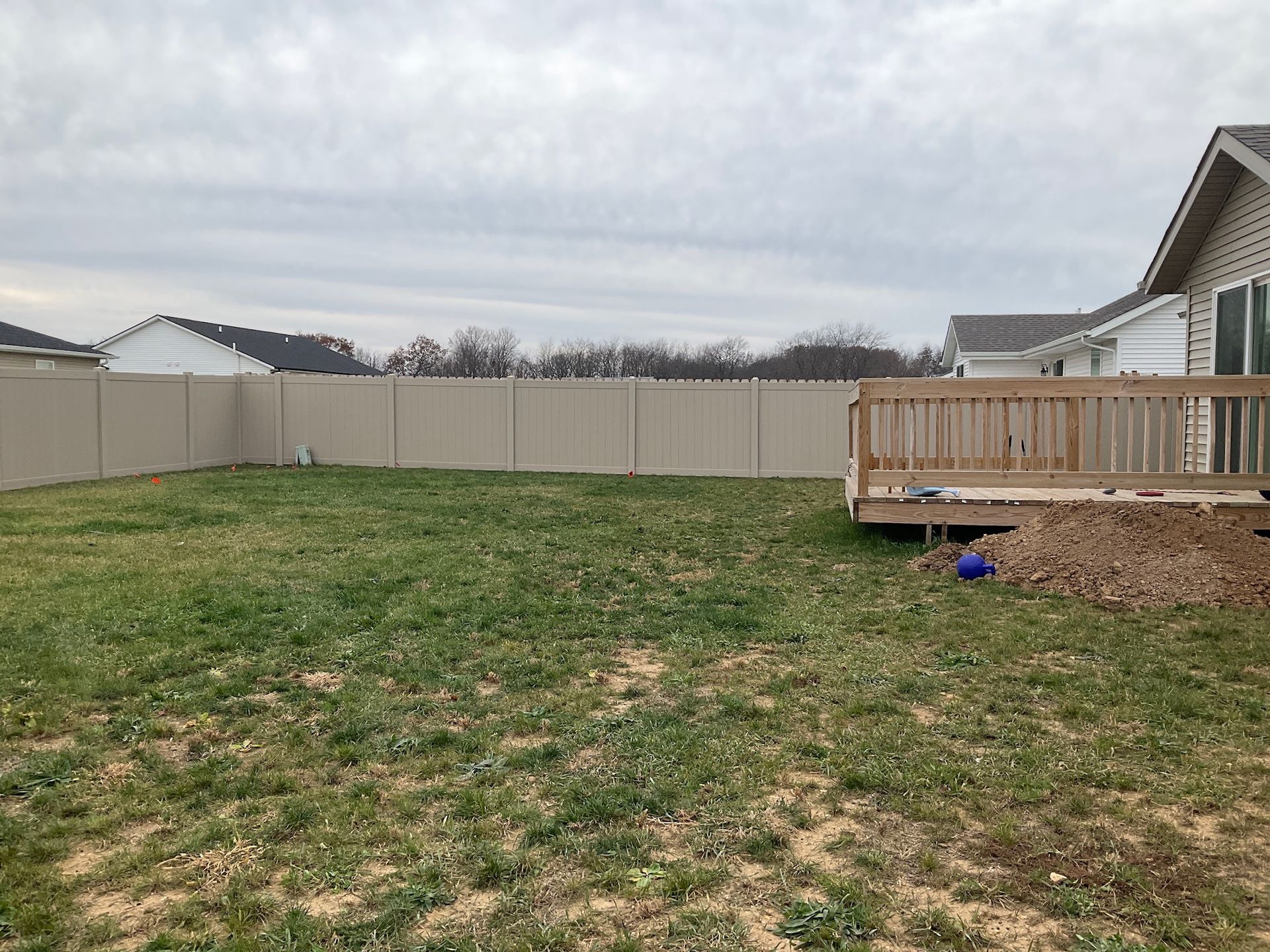 Backyard with tan fence, wood deck, and patchy green grass under an overcast sky.