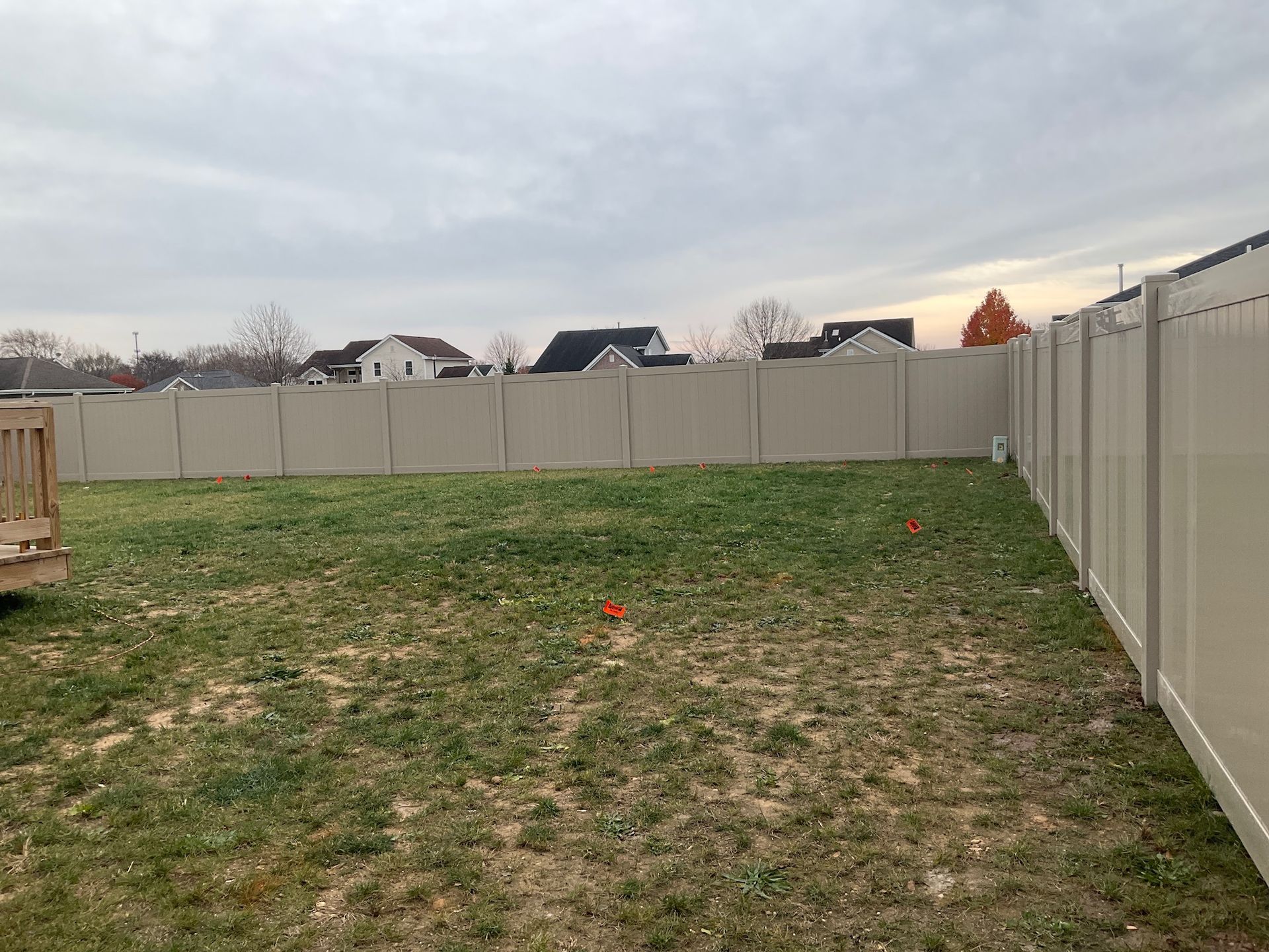 Beige vinyl fence encloses a backyard with patchy grass and a cloudy sky. Houses visible in the distance.