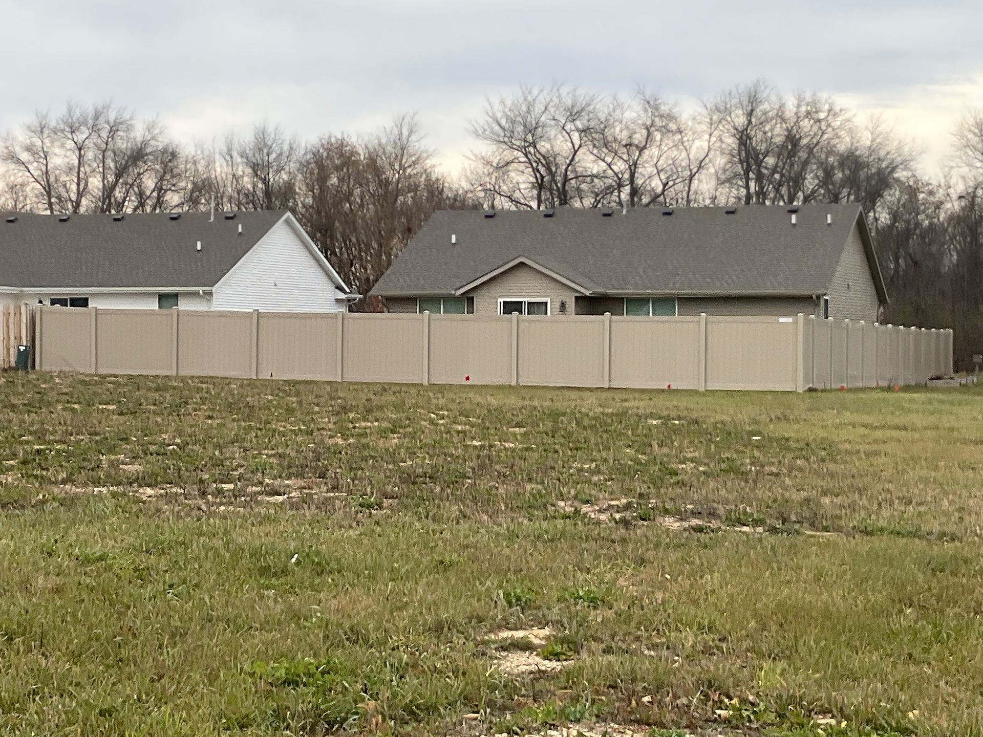 Tan privacy fence surrounding a house. Houses and trees are in the background. Overcast sky.