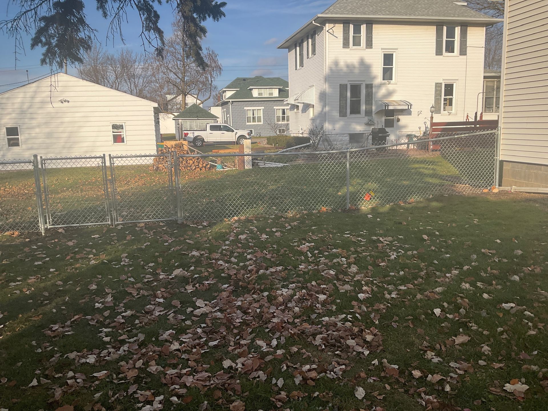 Backyard with fallen leaves, chain-link fence, and several houses on a sunny day.