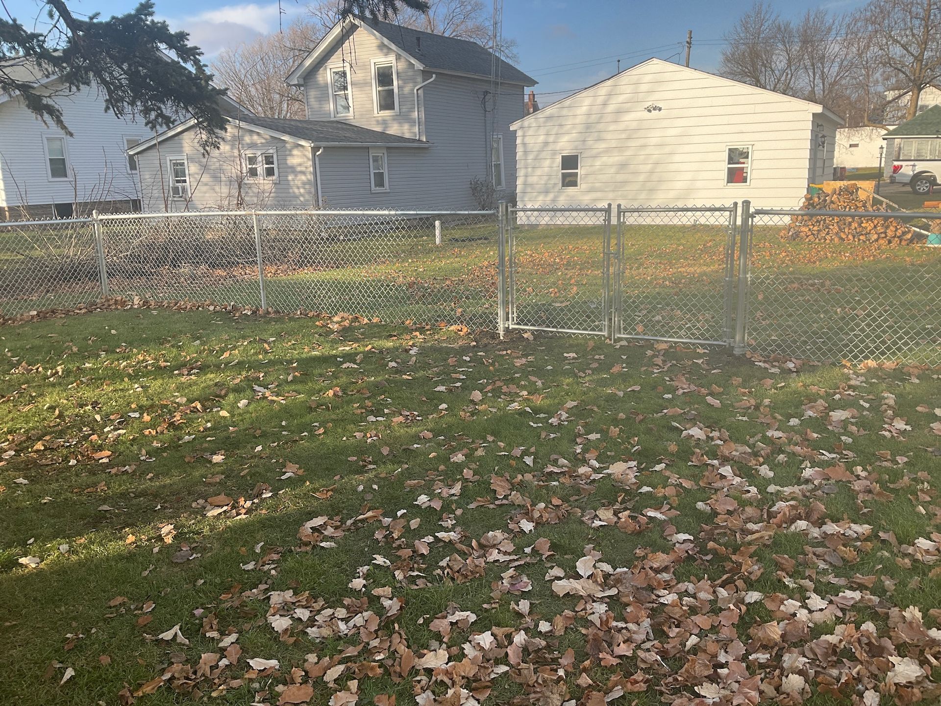 A fenced-in backyard with a house and detached garage. Leaves cover the grass.
