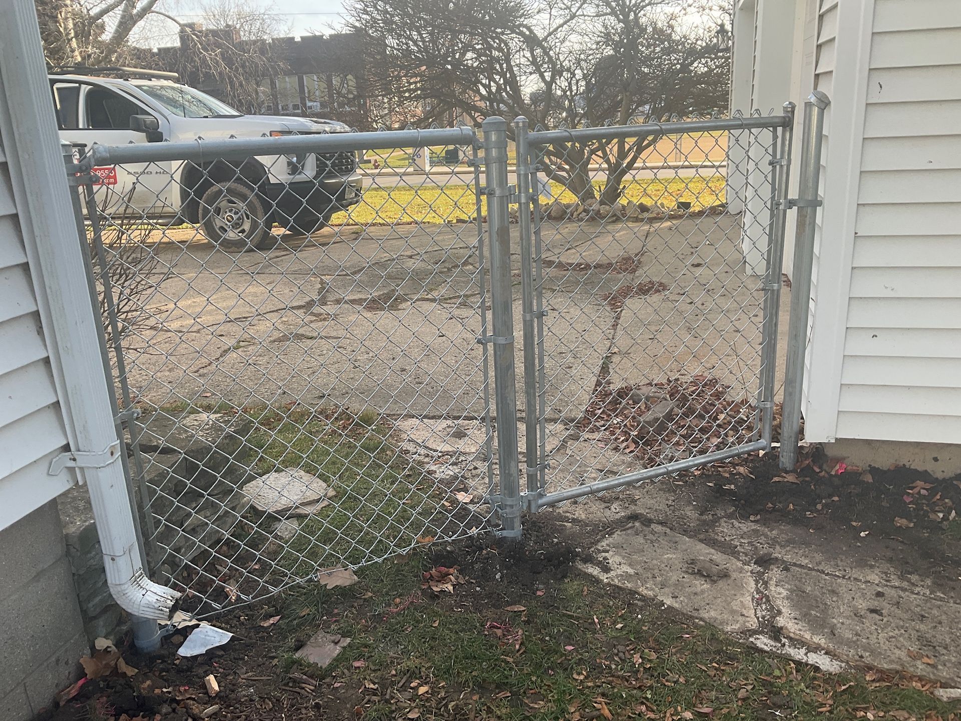 A chain link fence and gate next to a building and gutter. A vehicle is parked in the background.
