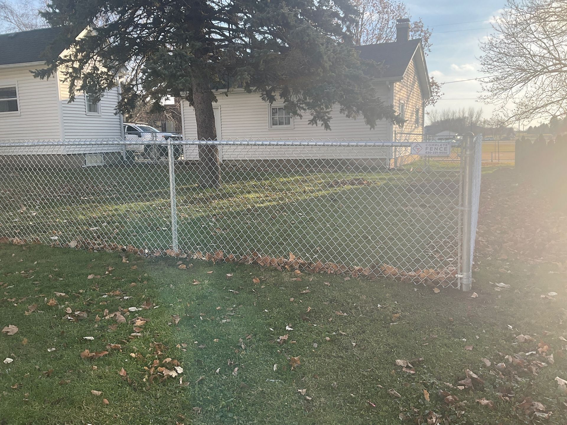 Chain-link fence in front yard with a house and tree in the background. Green grass and overcast sky.