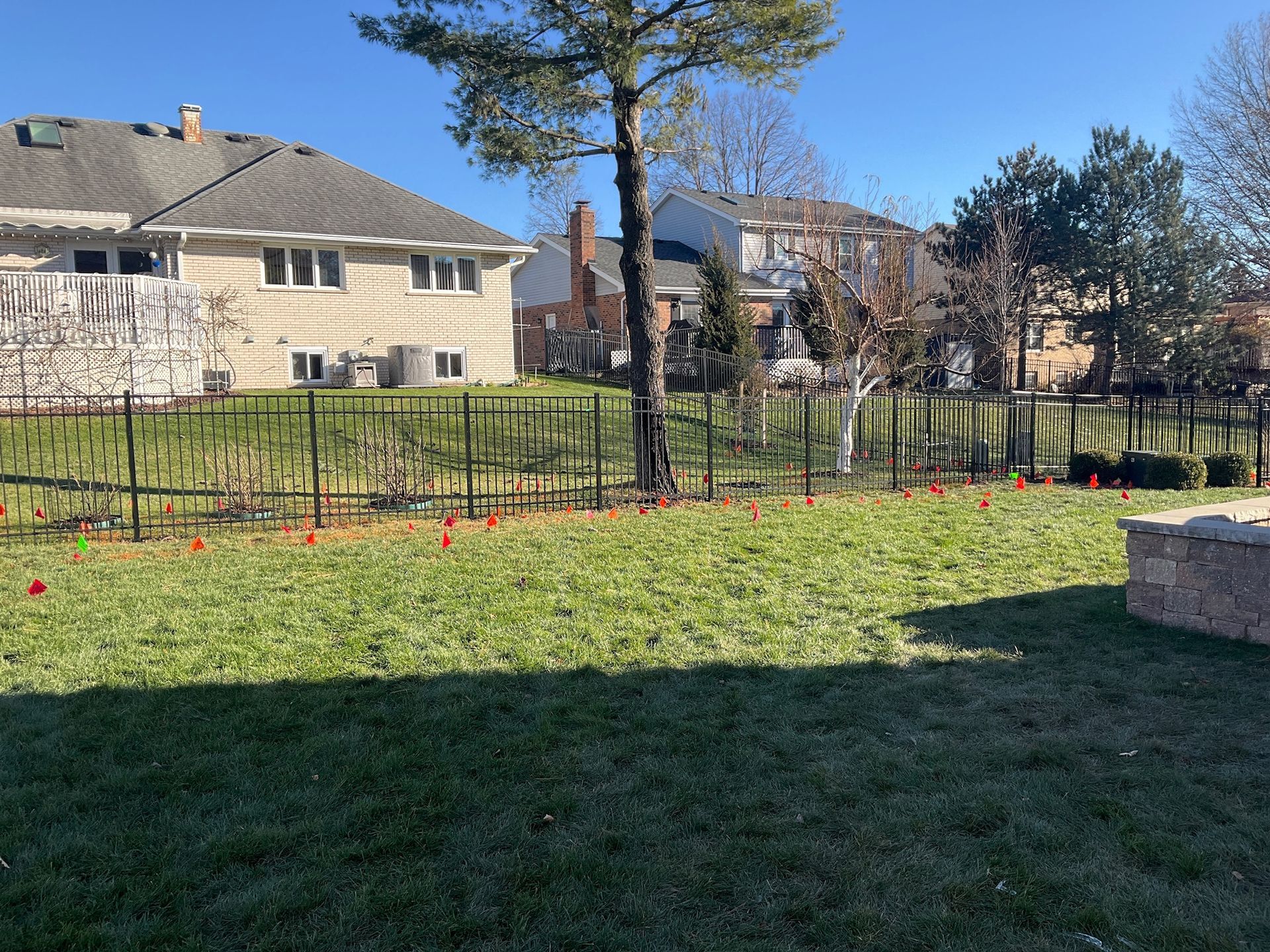 Backyard with green grass, black fence, and houses in the background on a sunny day. Orange flags mark the ground.
