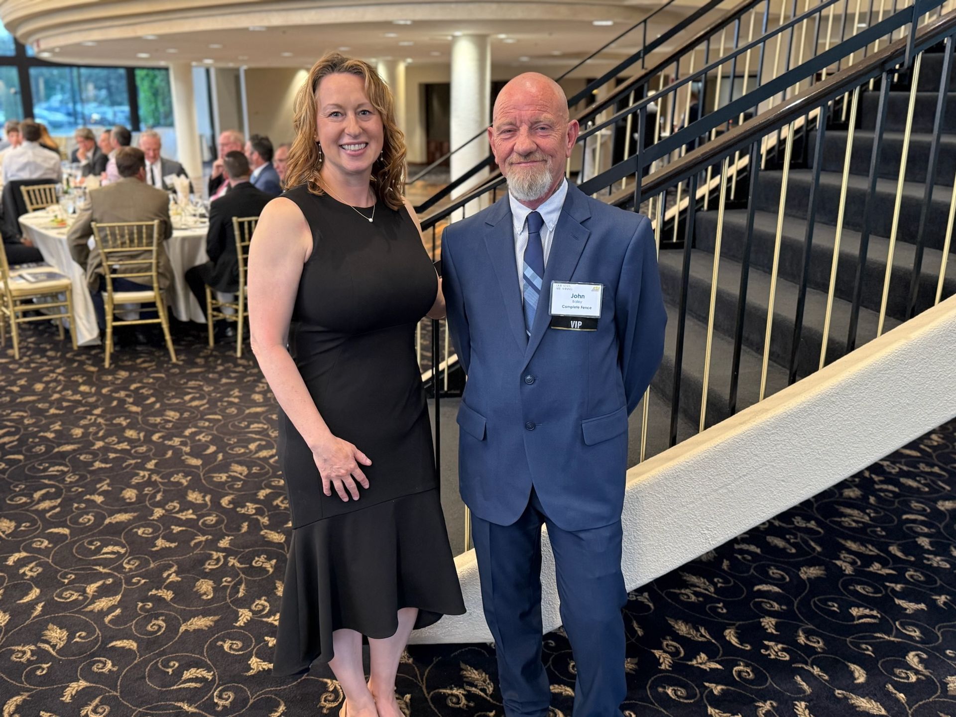 Woman in black dress and man in a suit stand near a staircase at an event. Other guests dine in the background.