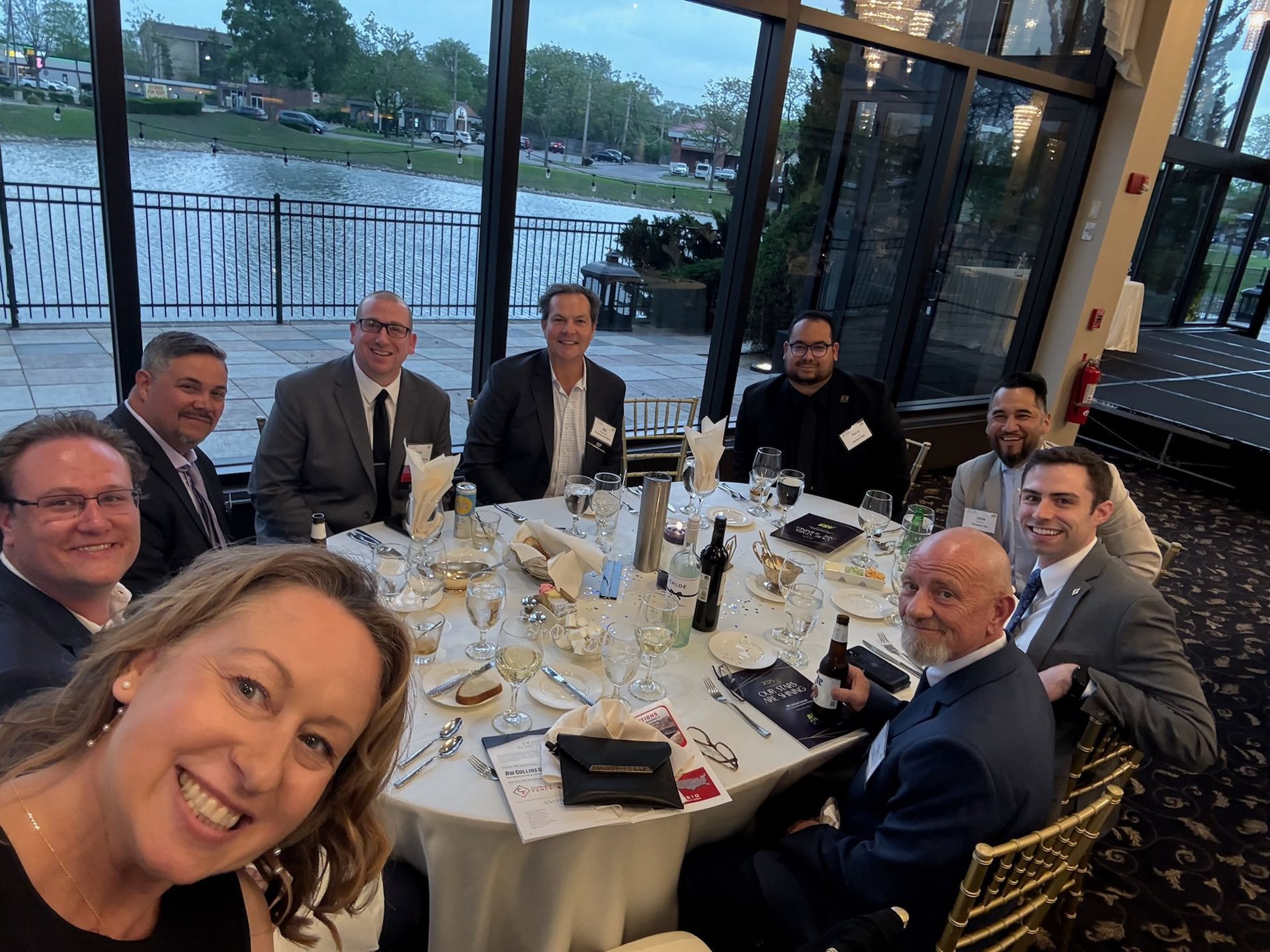 Group of people at a round table, smiling, with a waterfront view.