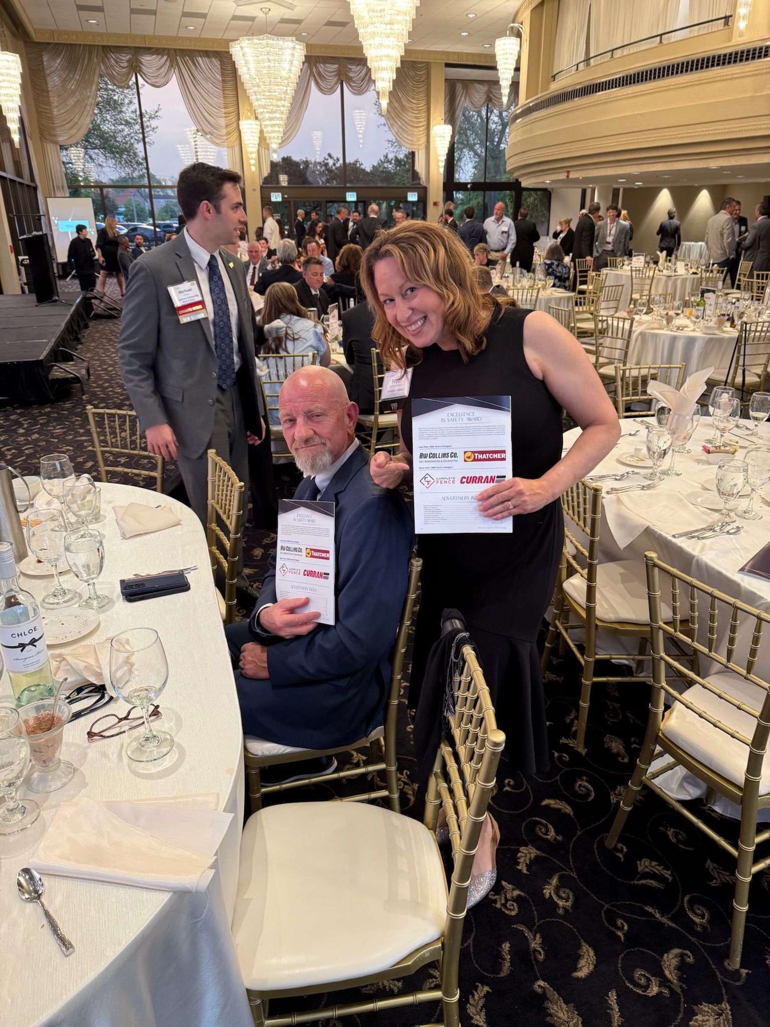 Two people holding papers smile at a gala, seated at a table. A man stands behind them.