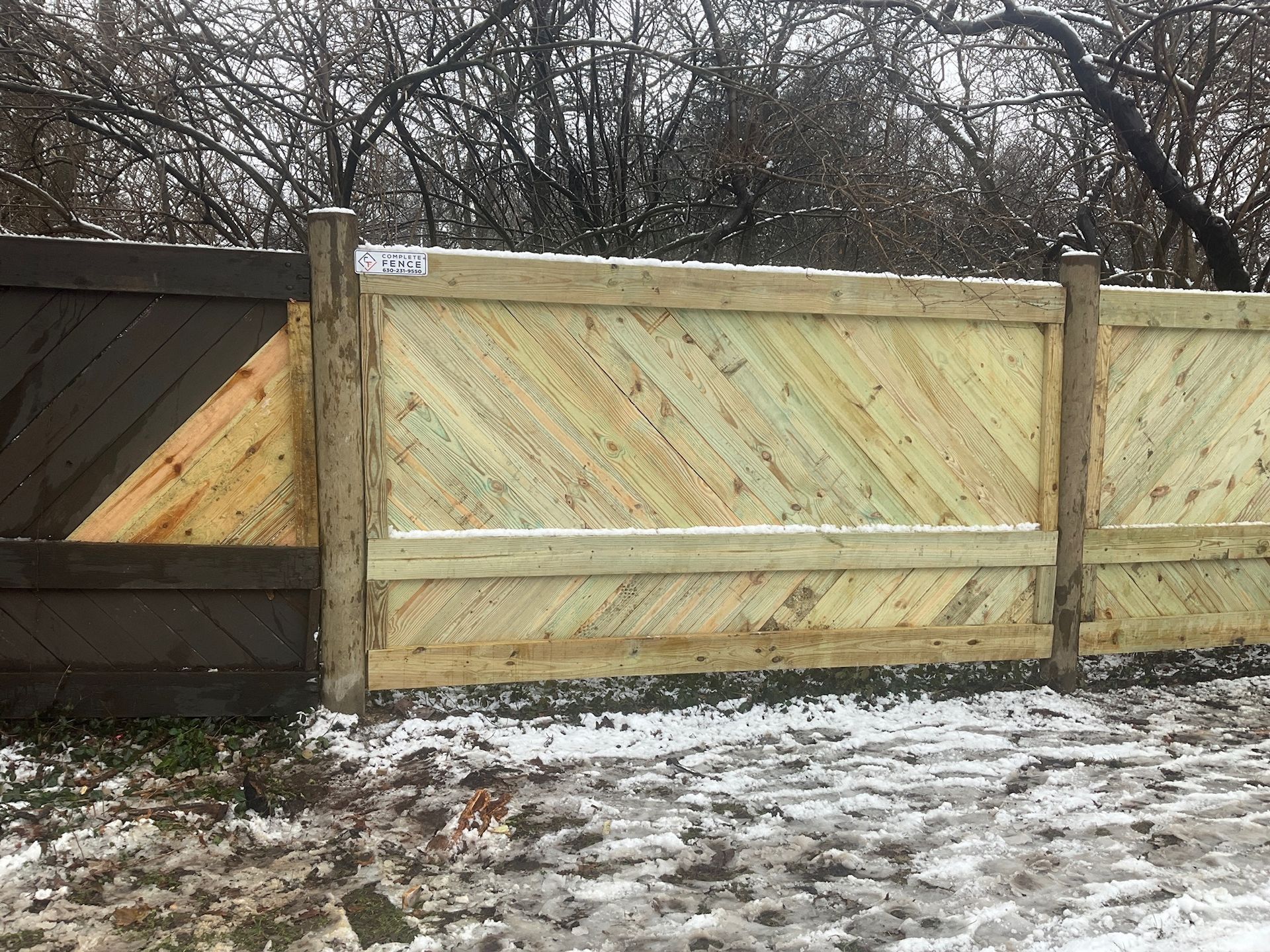 Wooden fence with a diagonal pattern, snow-covered ground, and trees in the background.