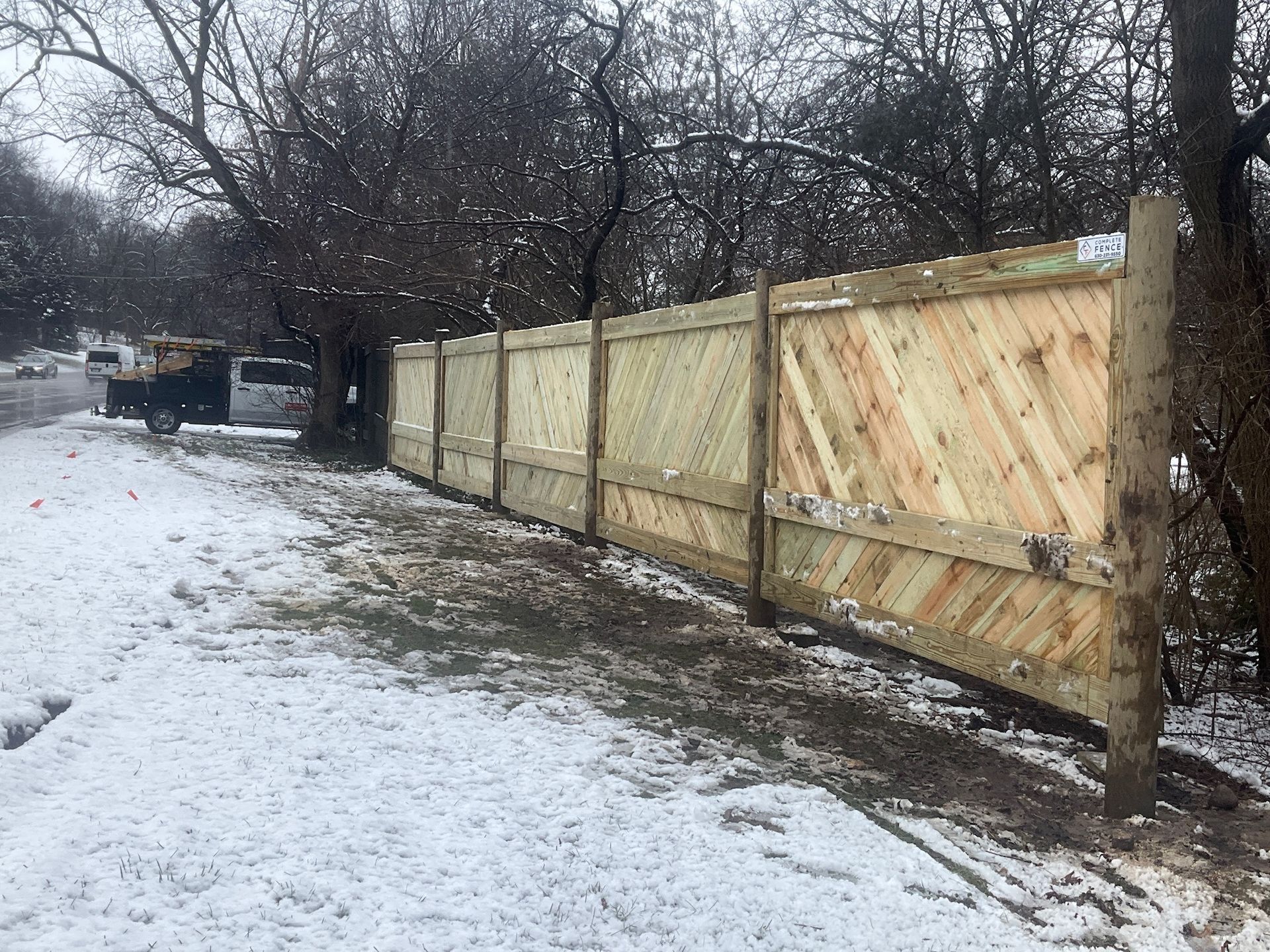 A wooden fence with diagonal pattern, covered in snow, along a road on a cloudy day.