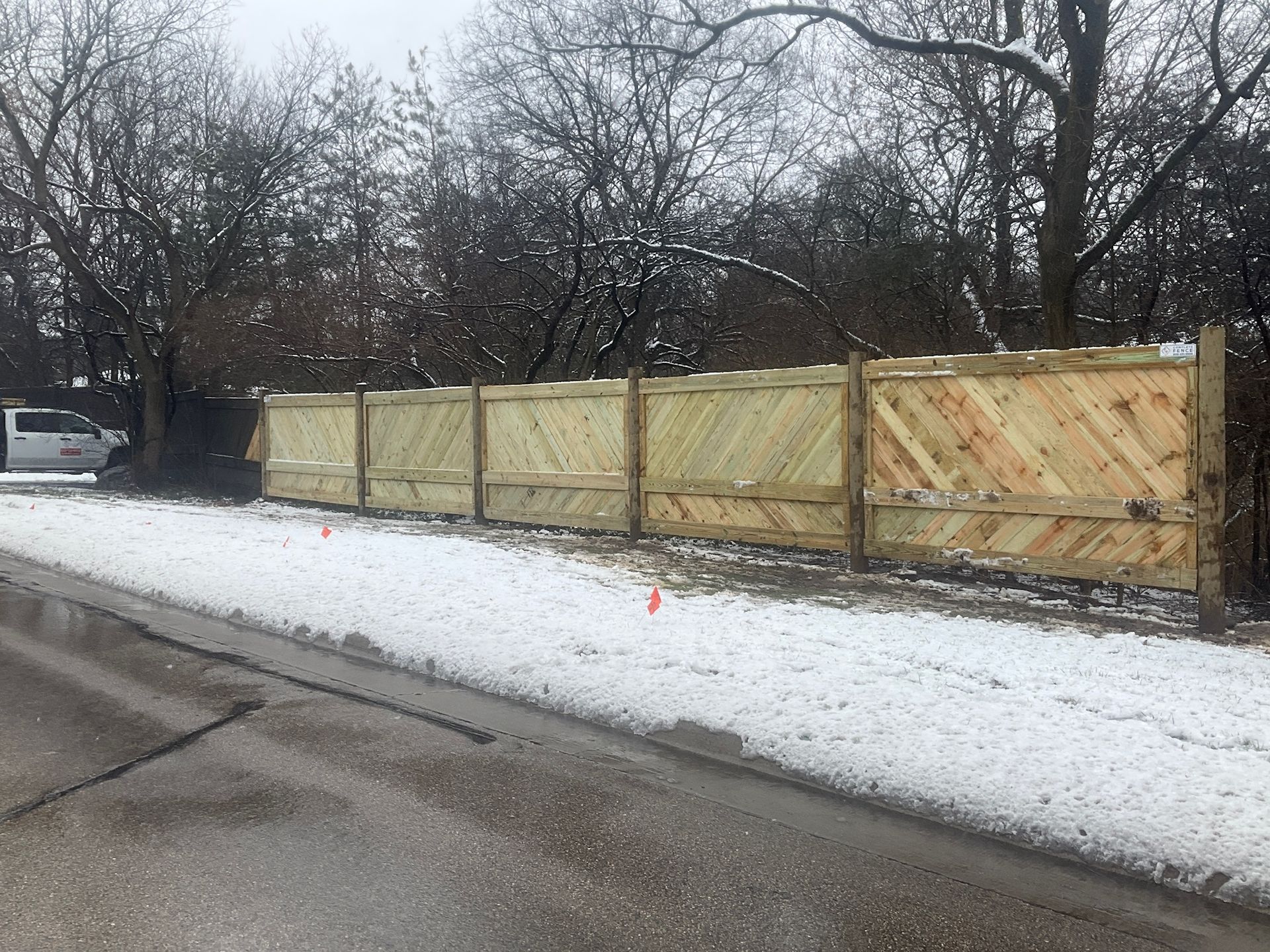 Wooden fence with diagonal paneling along snowy roadside. Bare trees in background, overcast sky.