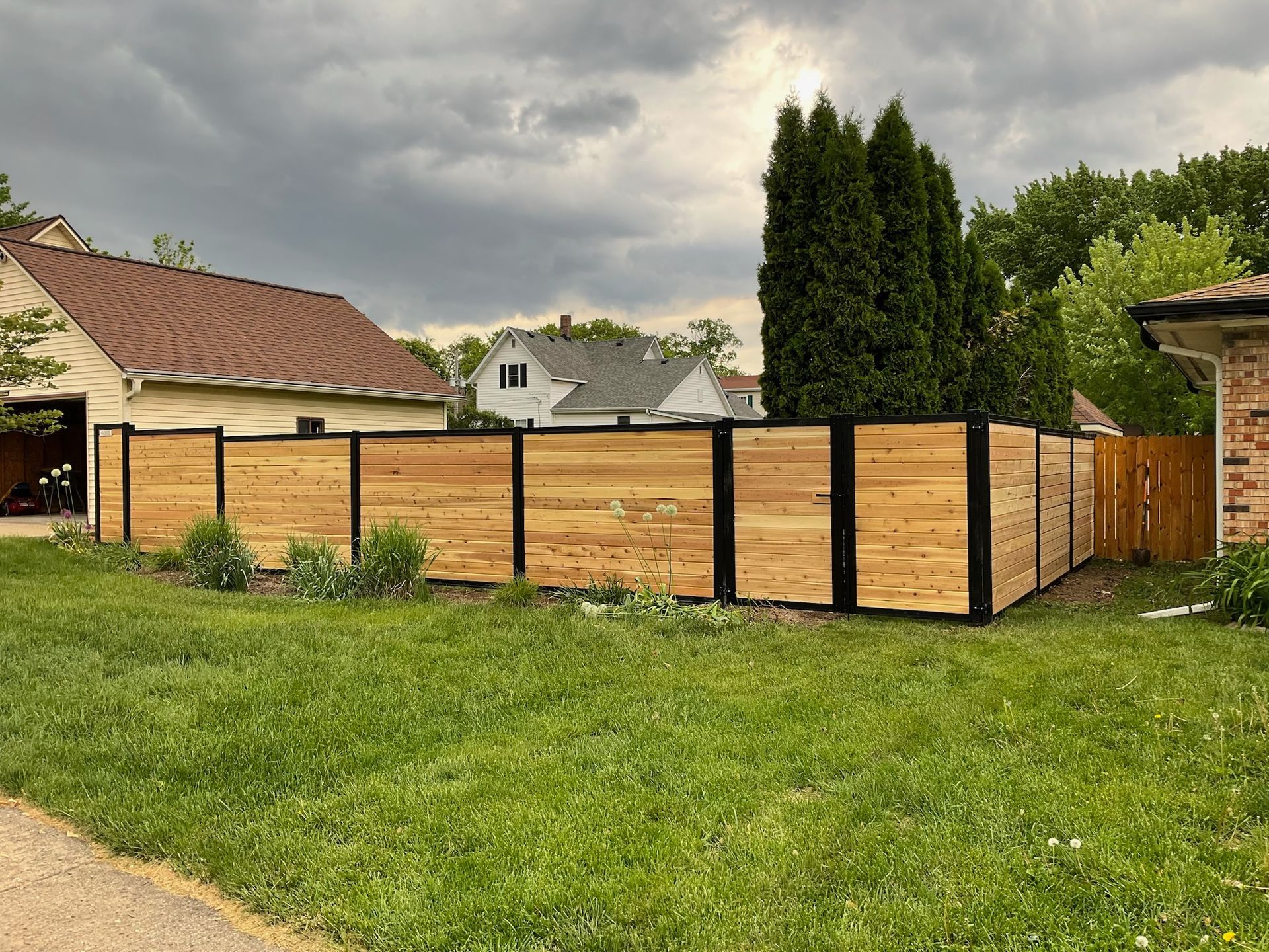 A wooden fence with black frames in front of a house, set in a grassy yard under a cloudy sky.