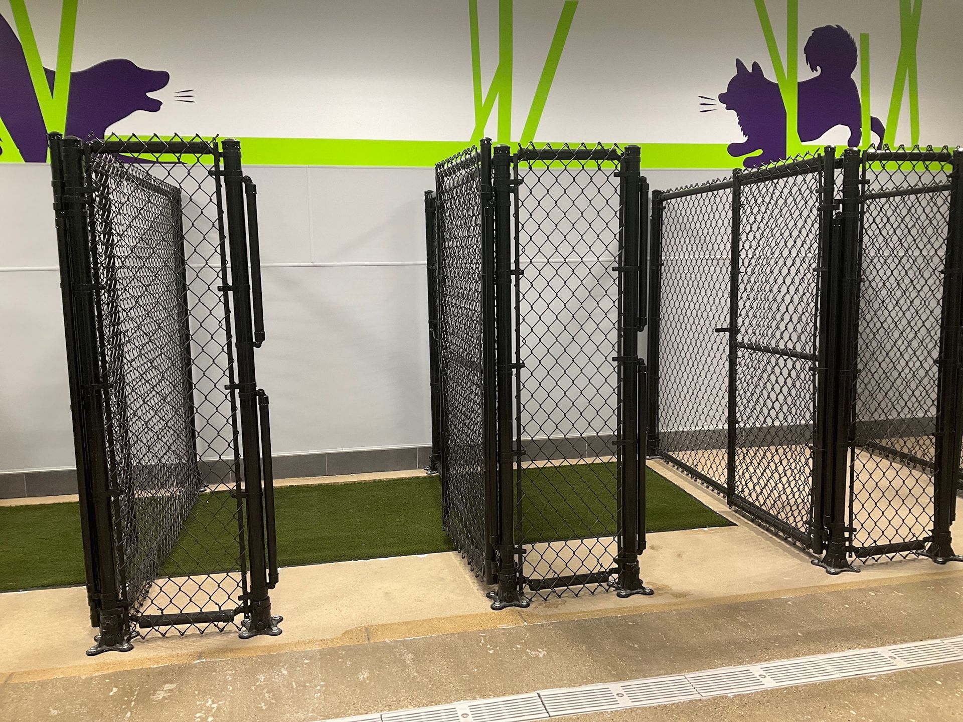 Three black chain-link fence enclosures with gates in a dog park with green turf and dog silhouettes on the wall.