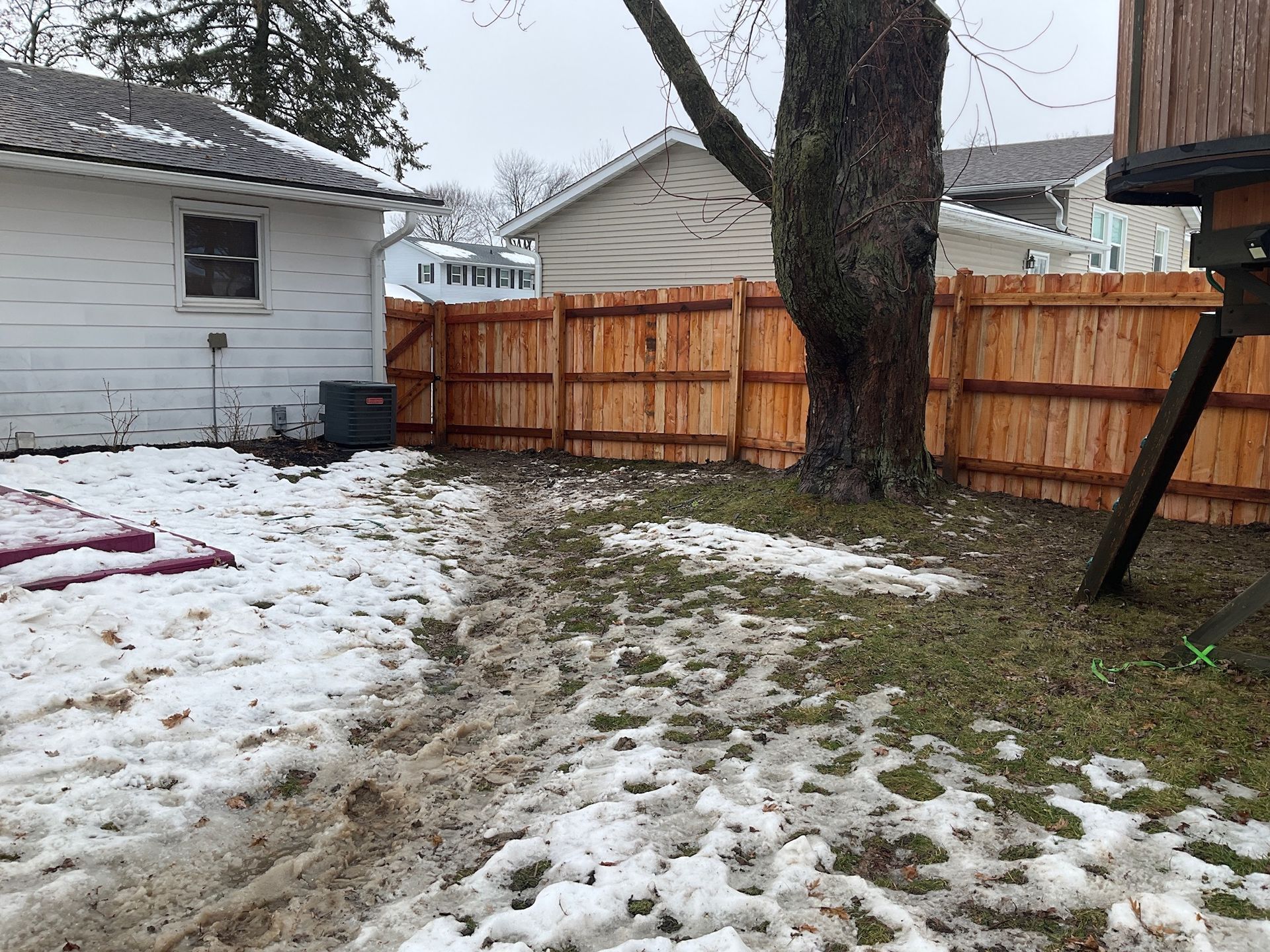 Backyard with patchy snow, wooden fence, tree, and small shed.