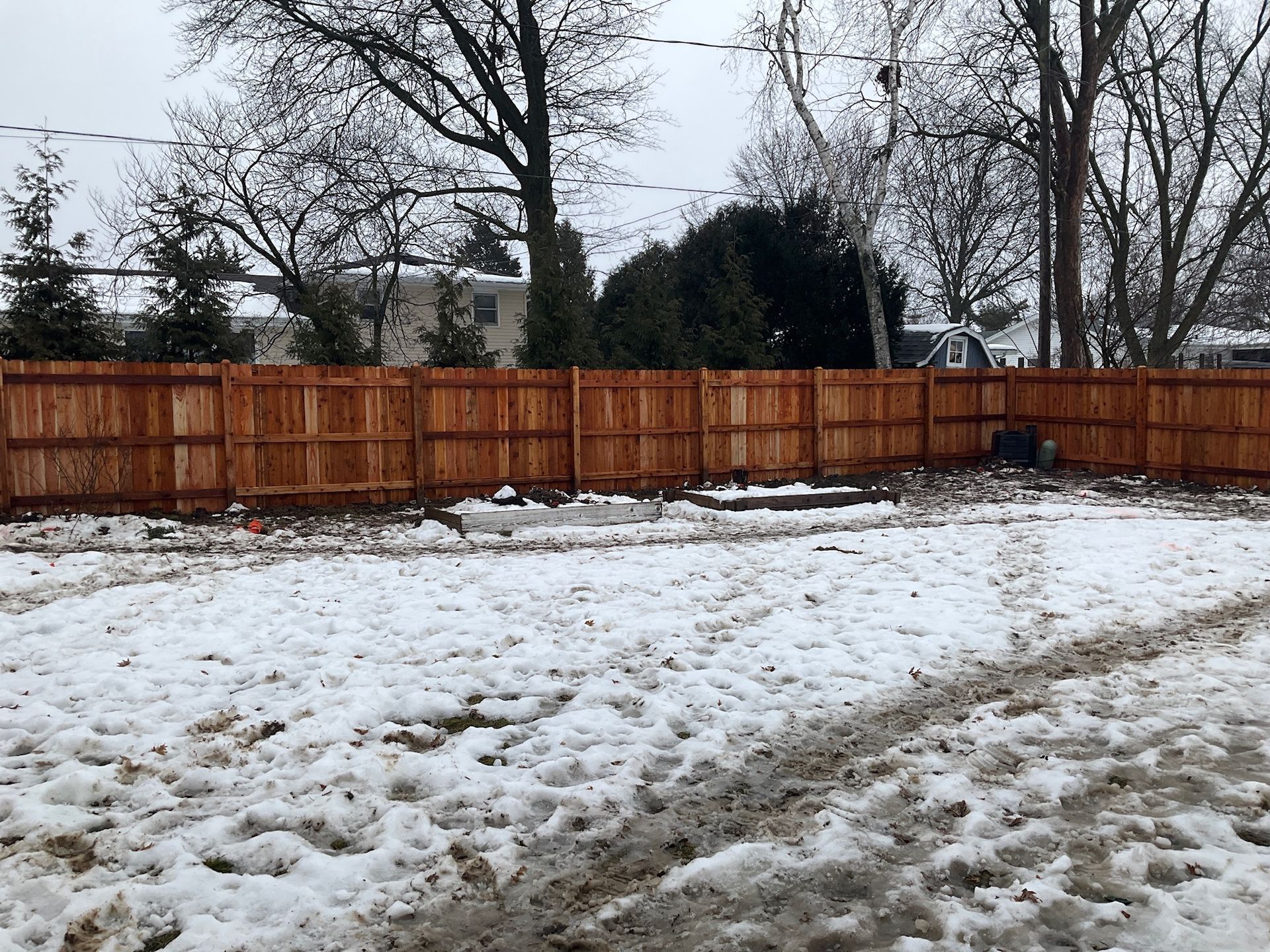 Snowy backyard with a wooden fence. Ground covered in snow, some bare patches.