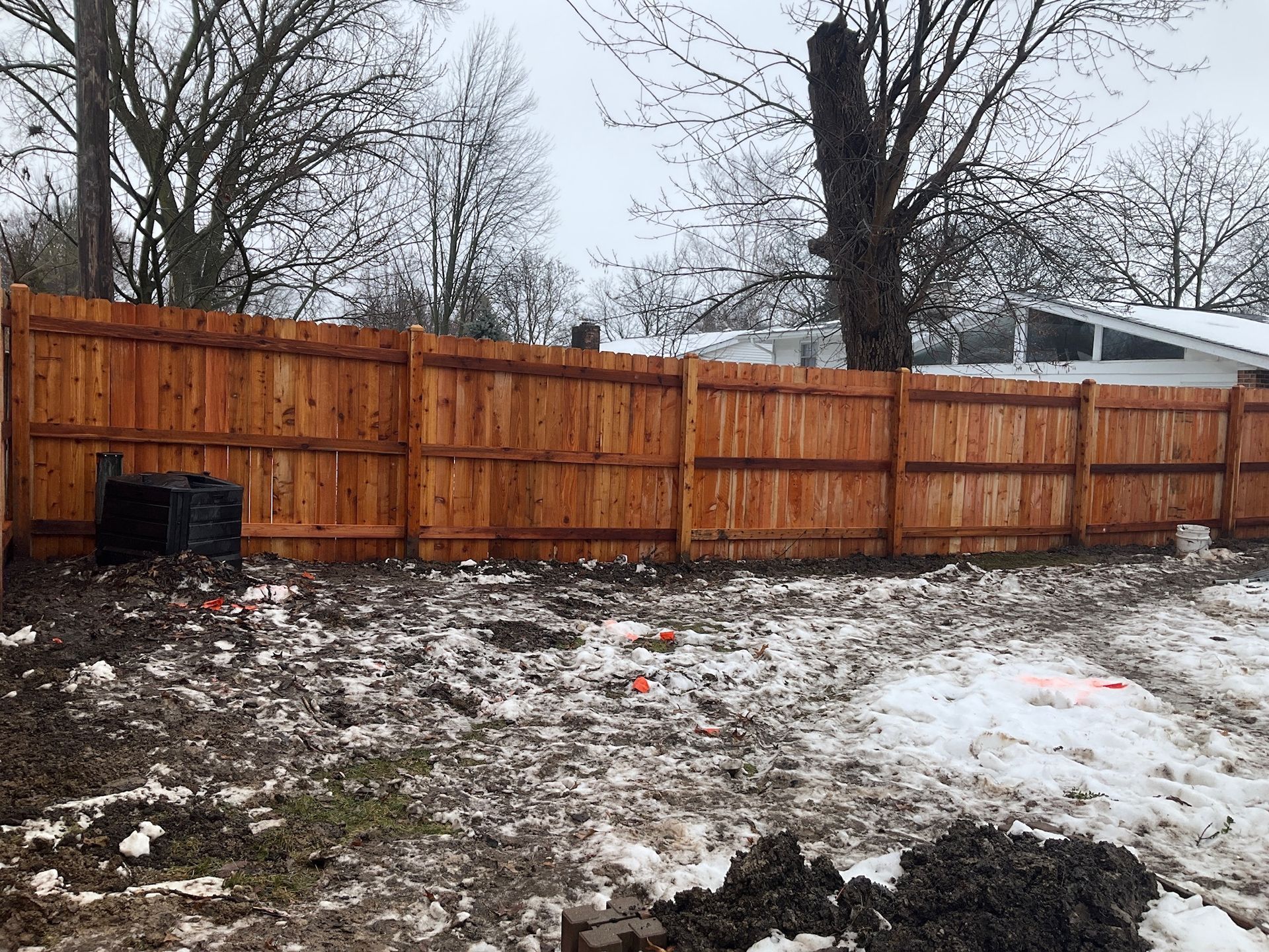 Wooden fence in a backyard with patchy snow on the ground; trees in the background.