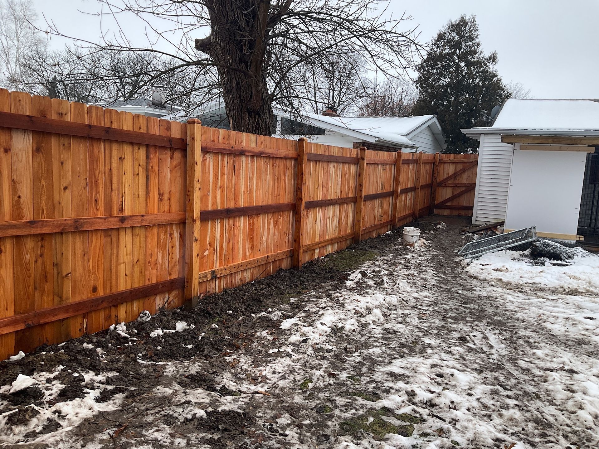 Wooden fence stained orange, running along a snow-covered backyard in winter.