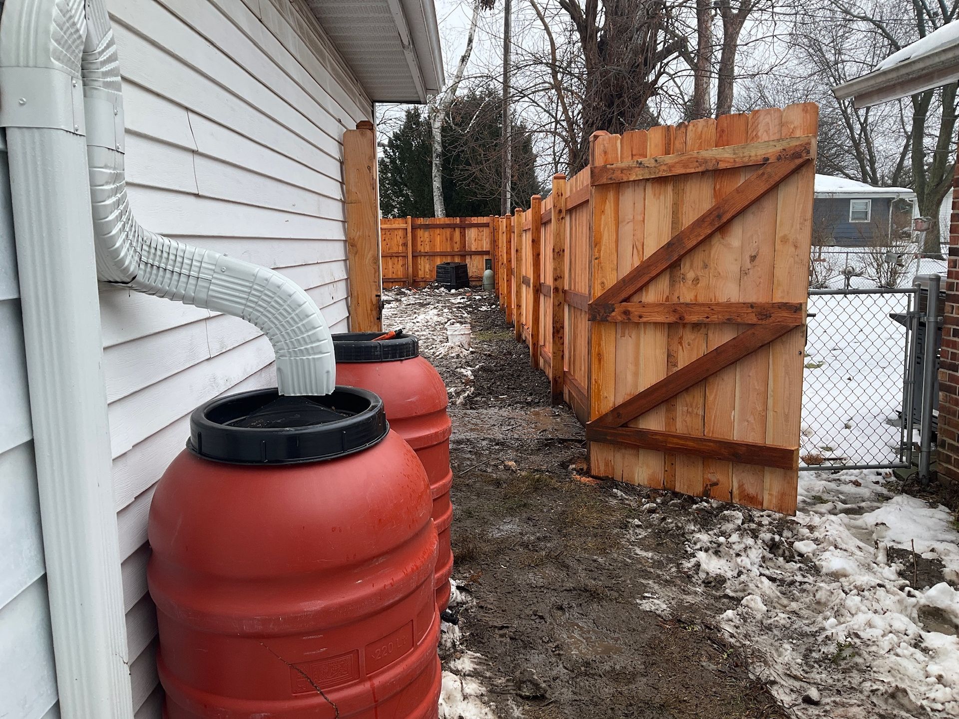 Two red rain barrels next to a white house, connected to a downspout, with a wooden fence in the background.