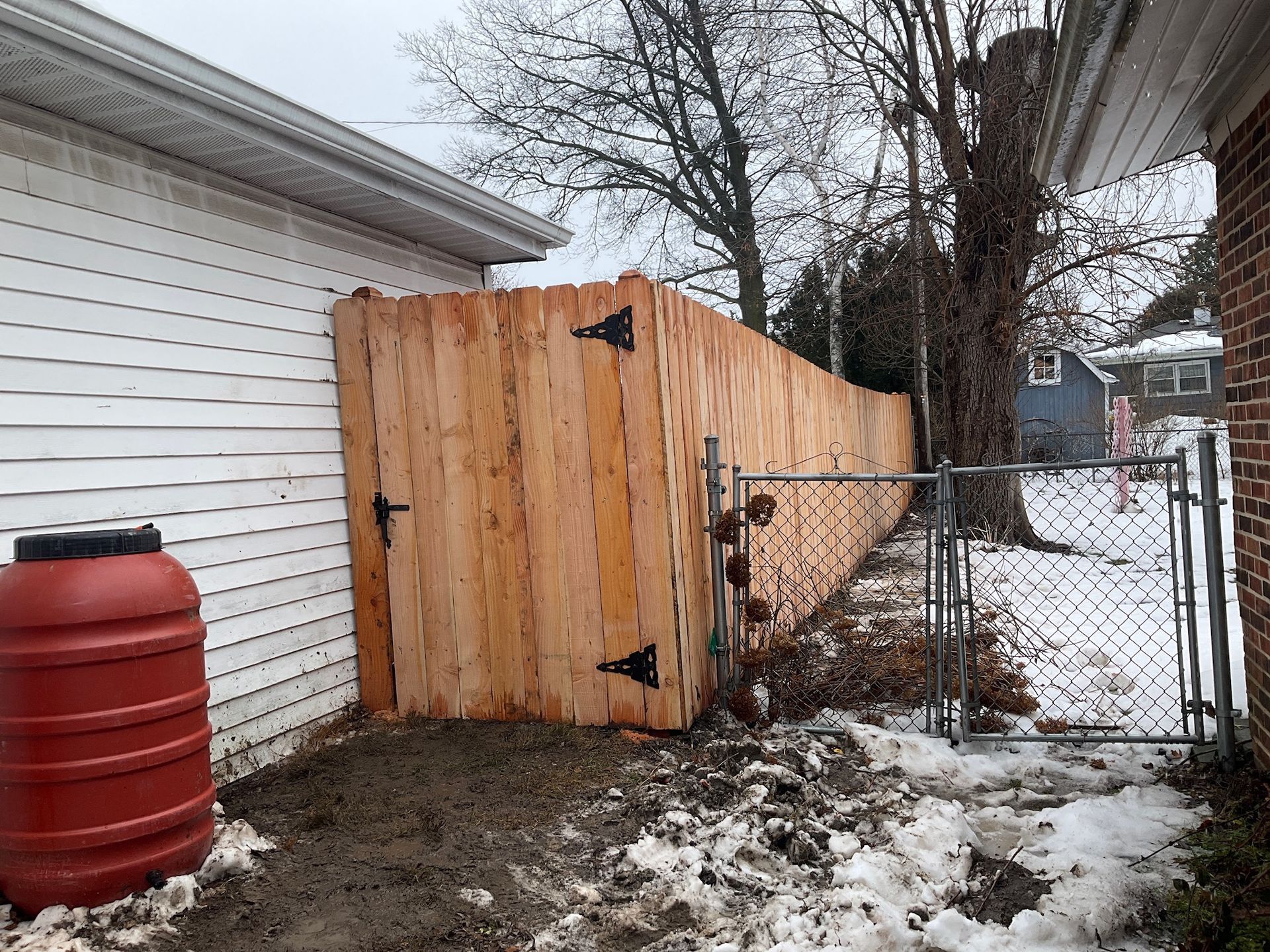 Wooden fence with a gate, snow on the ground, next to a white building and a chain-link fence.