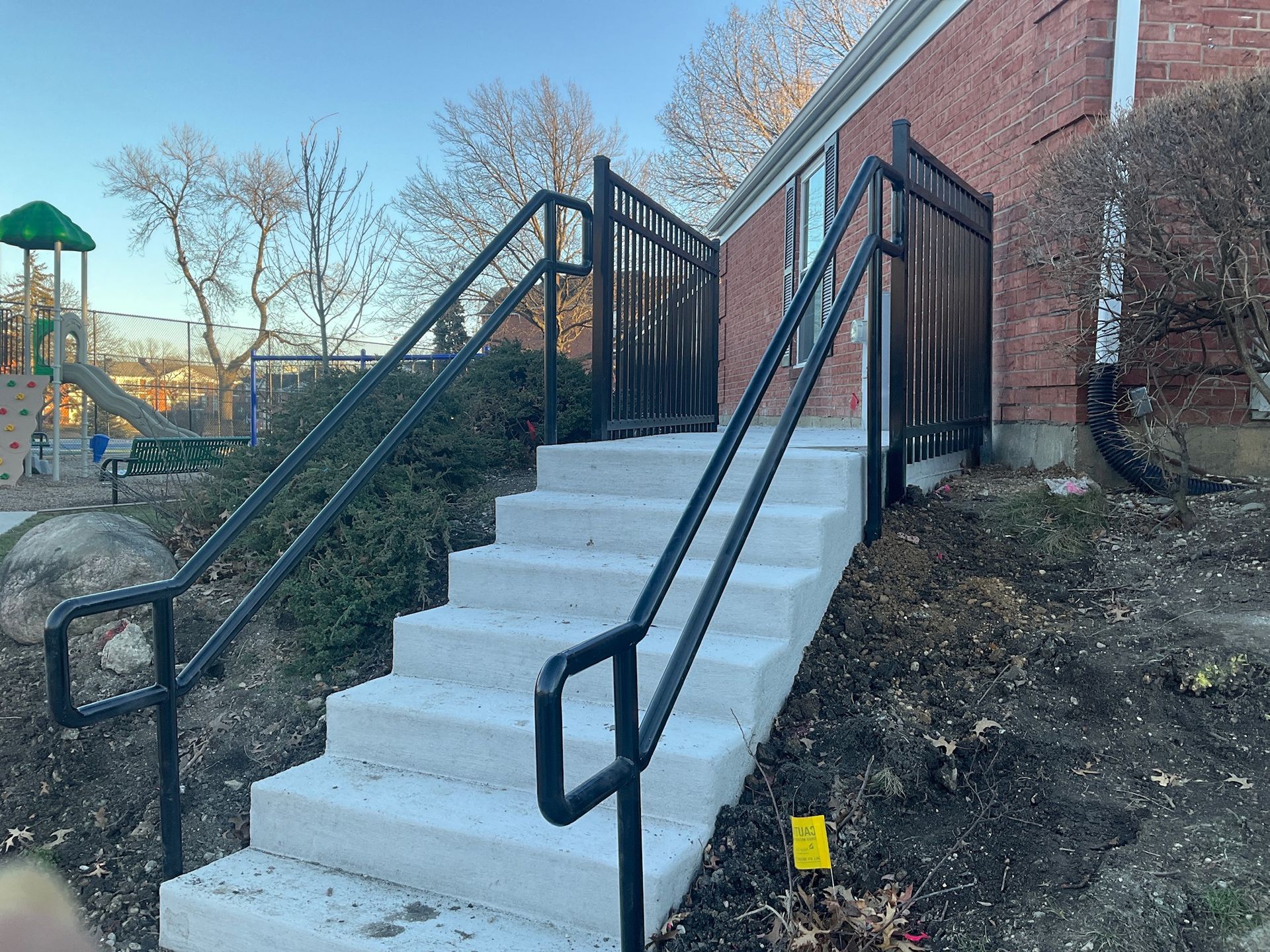 Concrete stairs with black handrails leading up to a brick building, alongside a fenced playground.