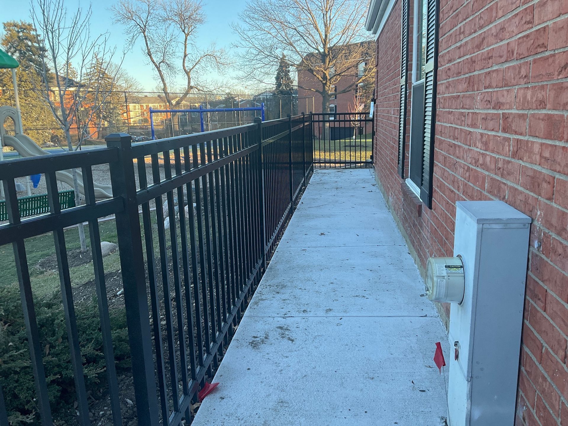 A concrete walkway next to a brick building and black metal fence. A playground is visible beyond the fence.